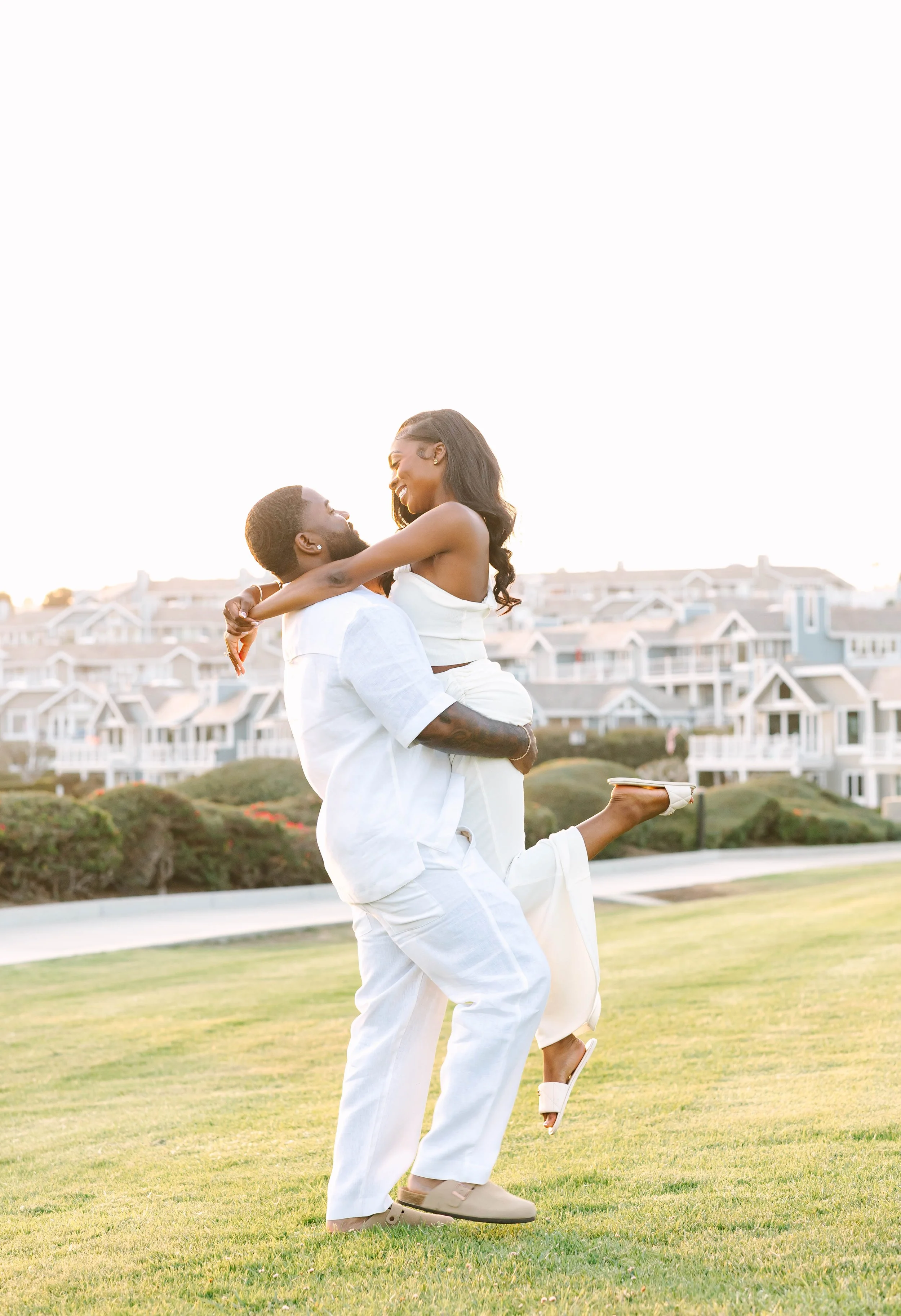 A couple in white clothing enjoying a moment outdoors, with one person lifting the other, in front of a background of houses or apartments on a grassy area during sunset.