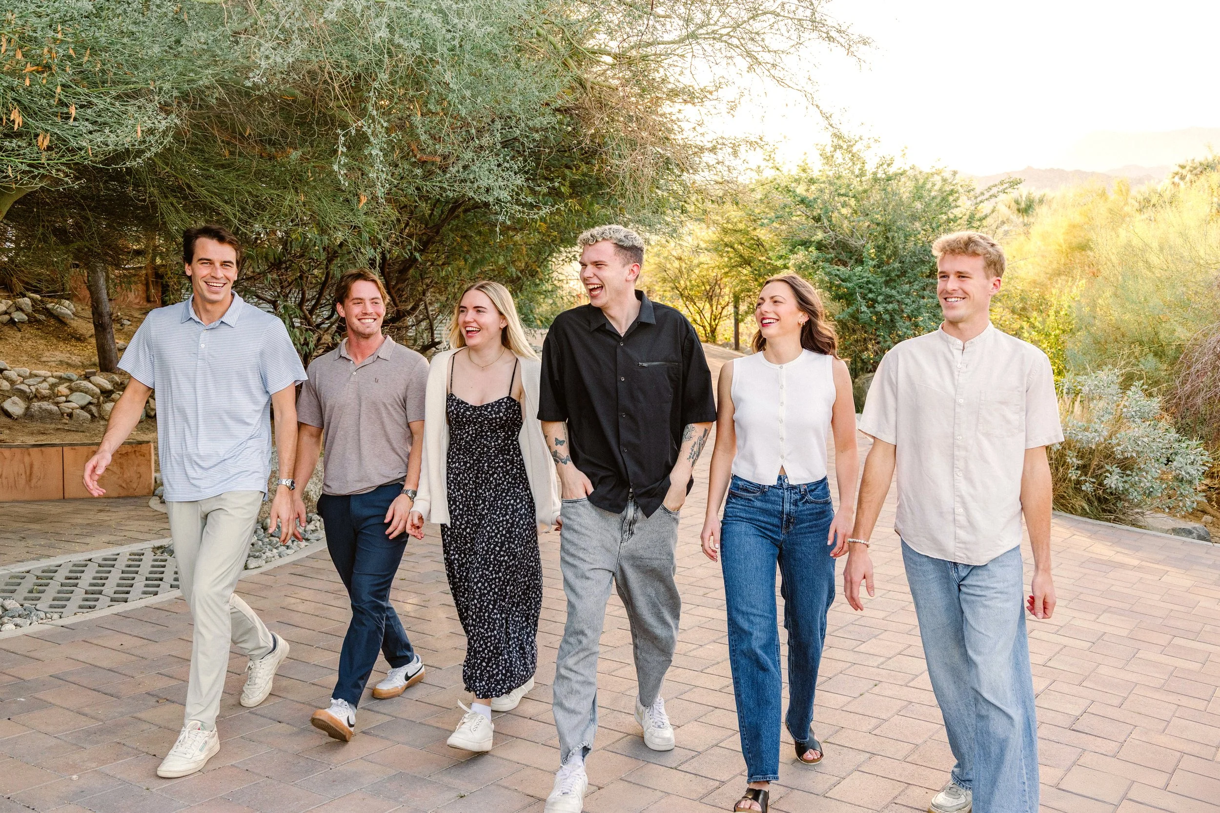 A group of six young adults walking outdoors on a paved path, smiling and laughing together, with trees and a sunny sky in the background.