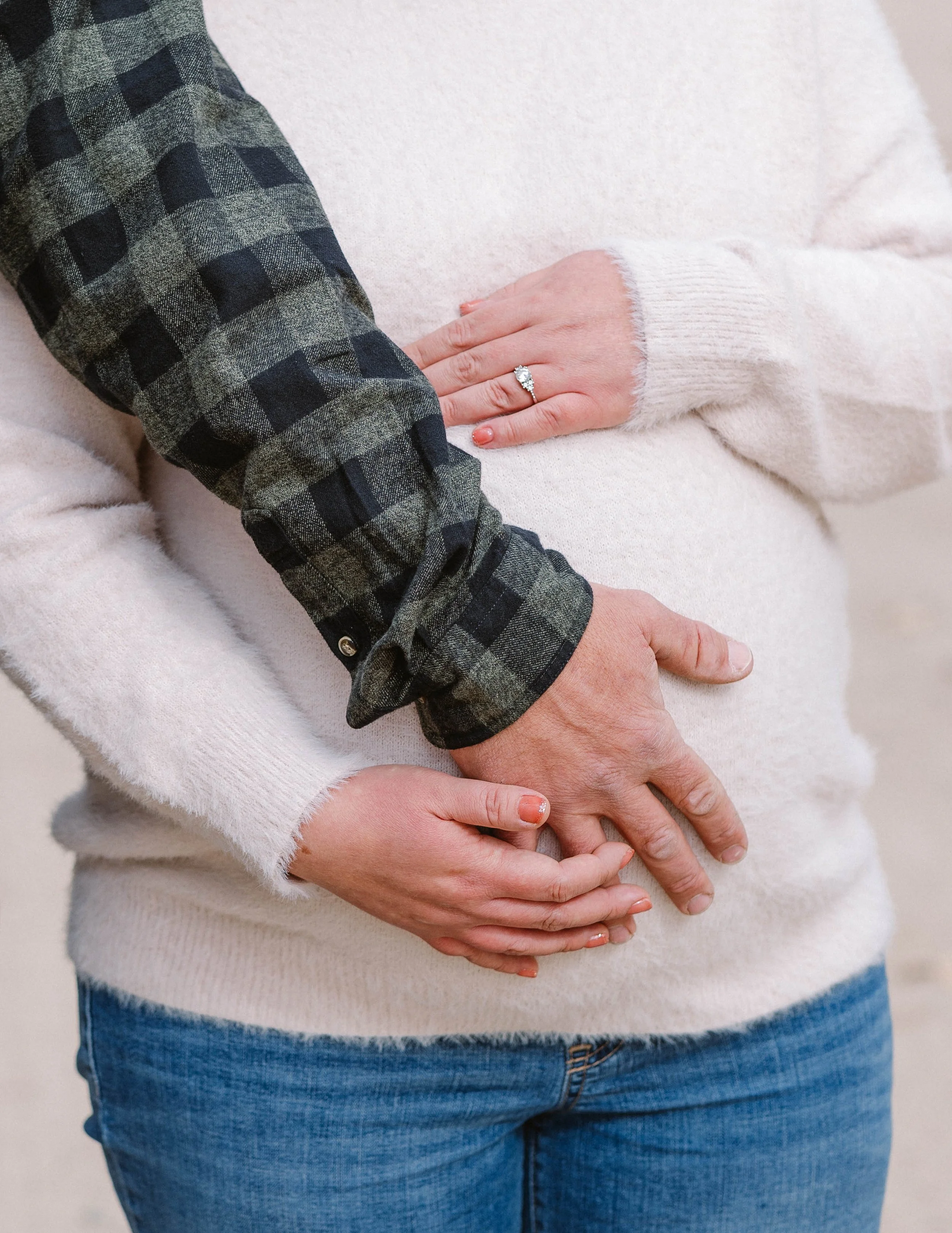 Close-up of a pregnant woman with an engagement ring on her finger, wearing a cream sweater and blue jeans, with someone else supporting her belly with hands.