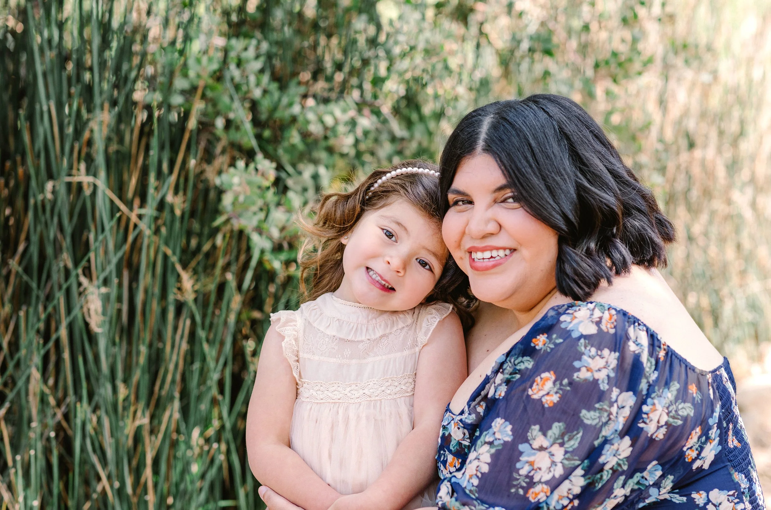 A woman and a young girl smiling and hugging outdoors near green bushes, with the woman wearing a blue floral dress and the girl in a light pink dress with lace details.