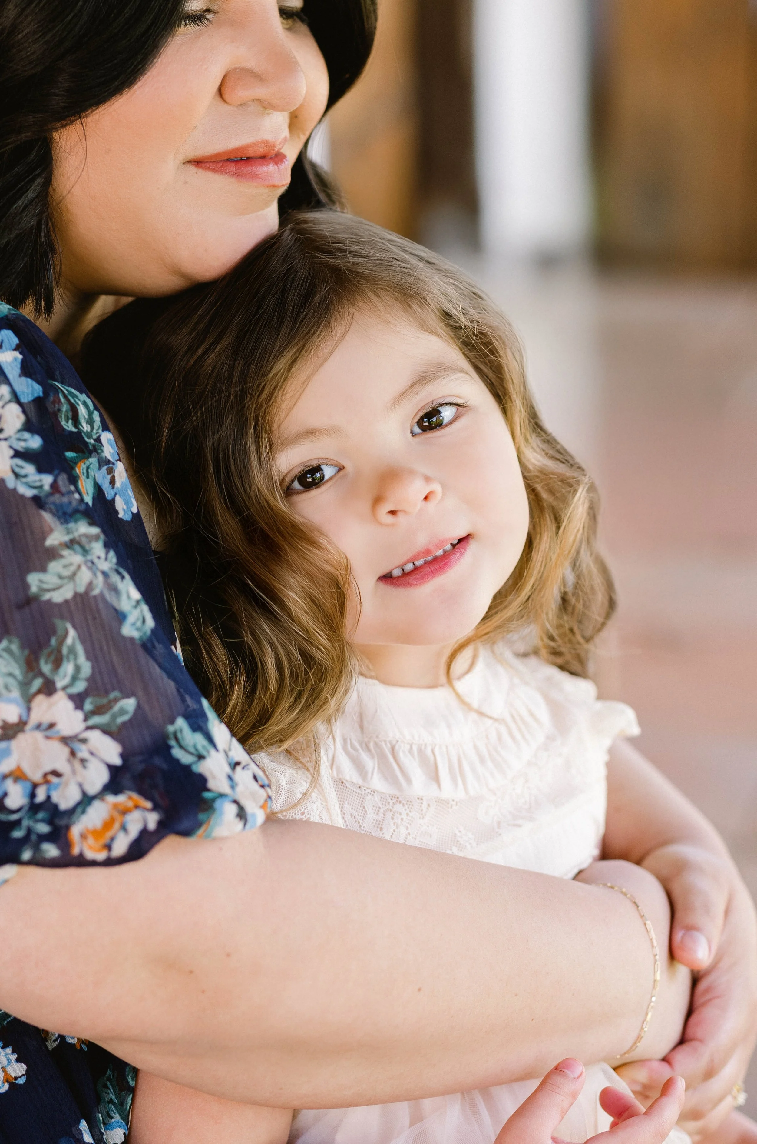 A woman holding a young girl close. The girl has curly hair and is looking at the camera with a gentle smile.