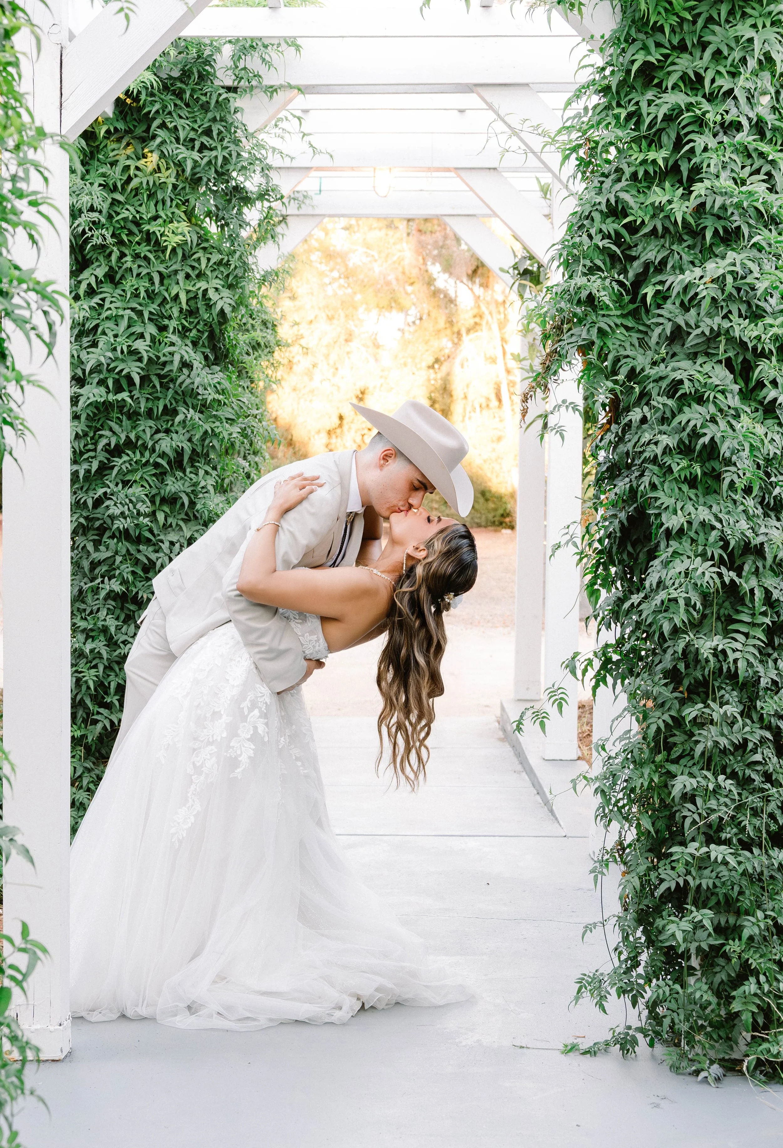 A bride and groom share a kiss and dip during their wedding photos in a lush, green, outdoor setting.