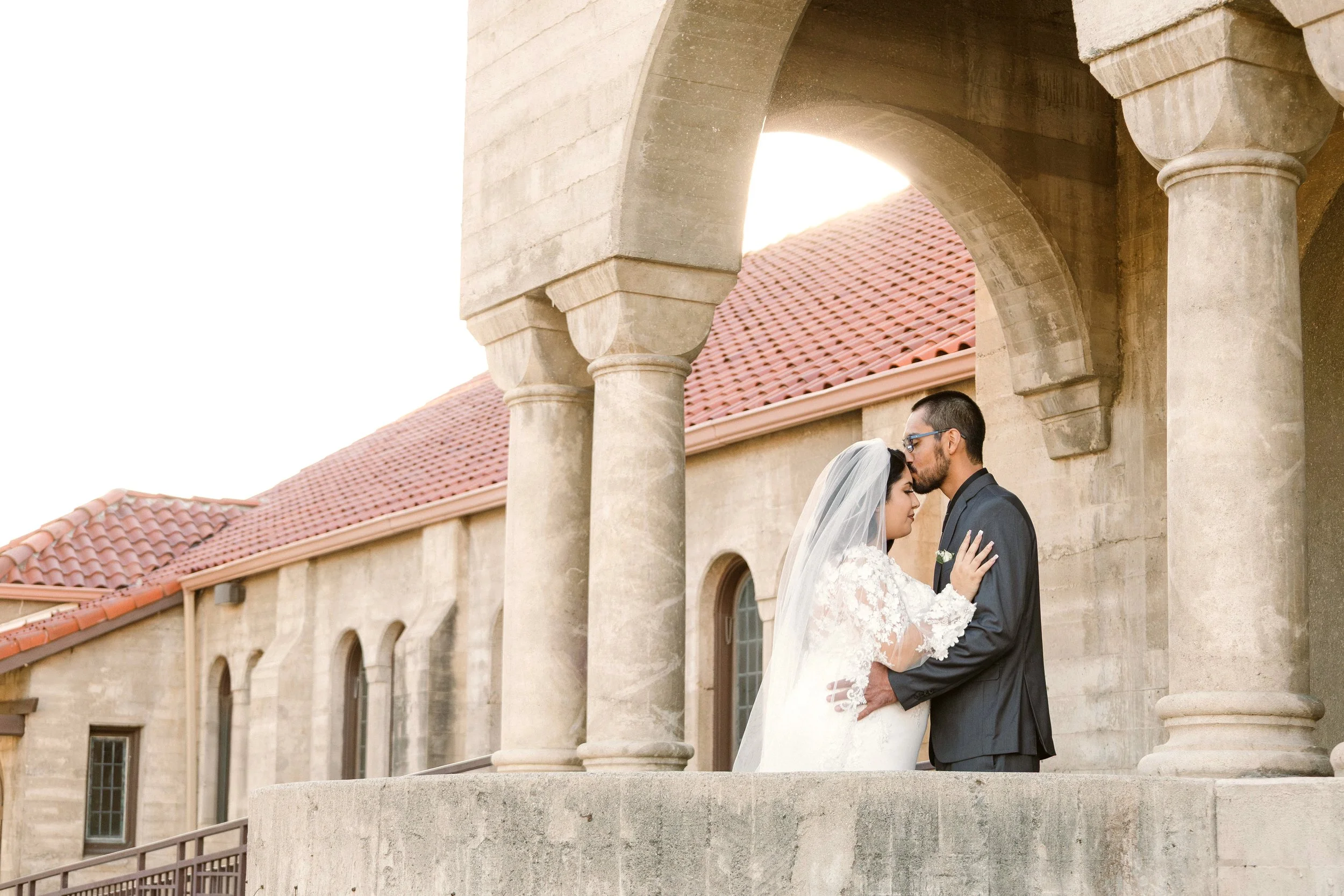 Couple in wedding attire standing under stone arches on a balcony, with a building with a red-tiled roof in the background.