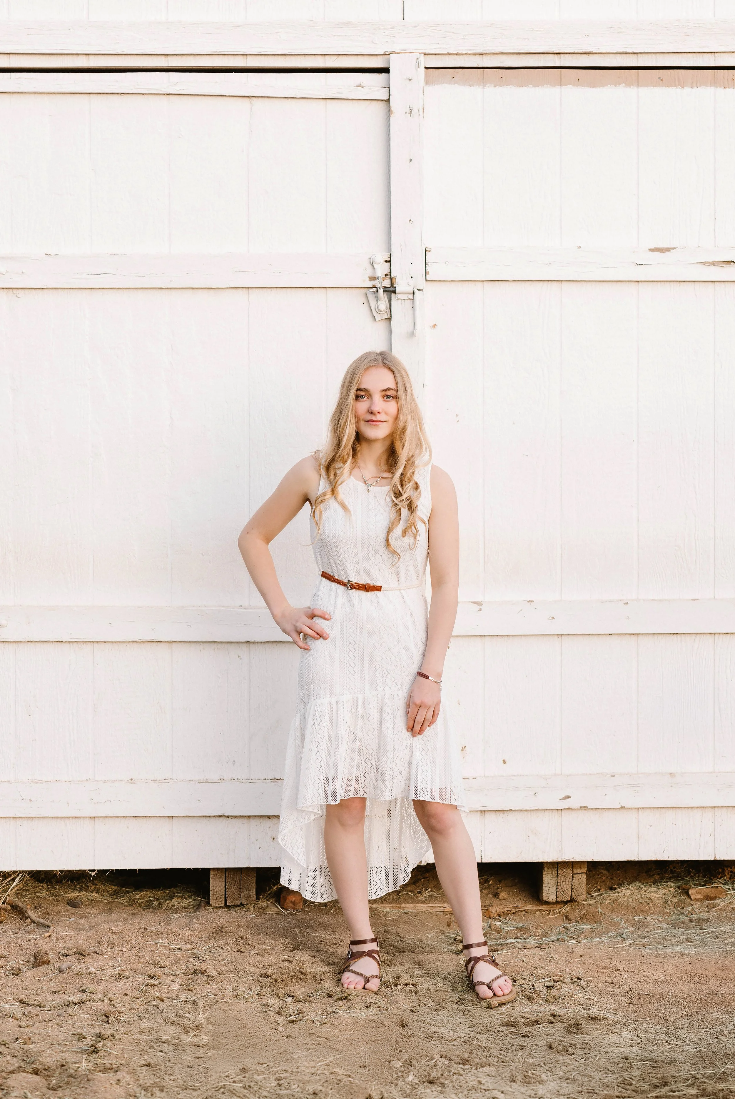 A young woman with long blonde hair wearing a white sleeveless lace dress, brown belt, and brown sandals standing in front of a white wooden barn door with dirt ground.