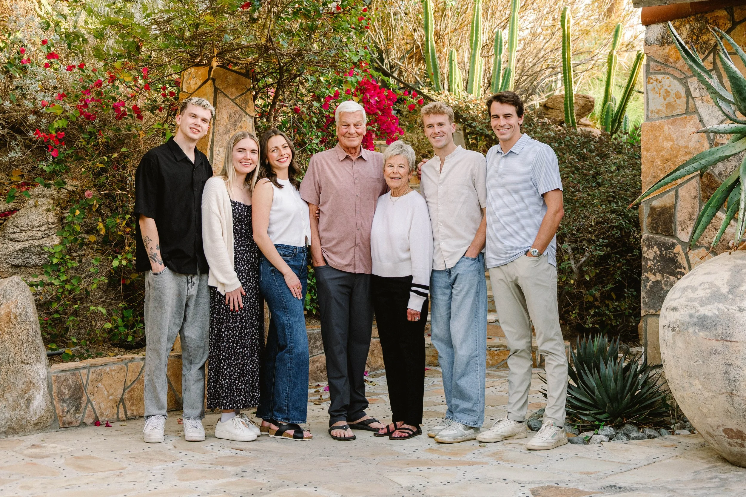 A multi-generational family of seven standing together outdoors in front of a lush garden with green plants, cacti, and vibrant pink flowers, smiling at the camera.