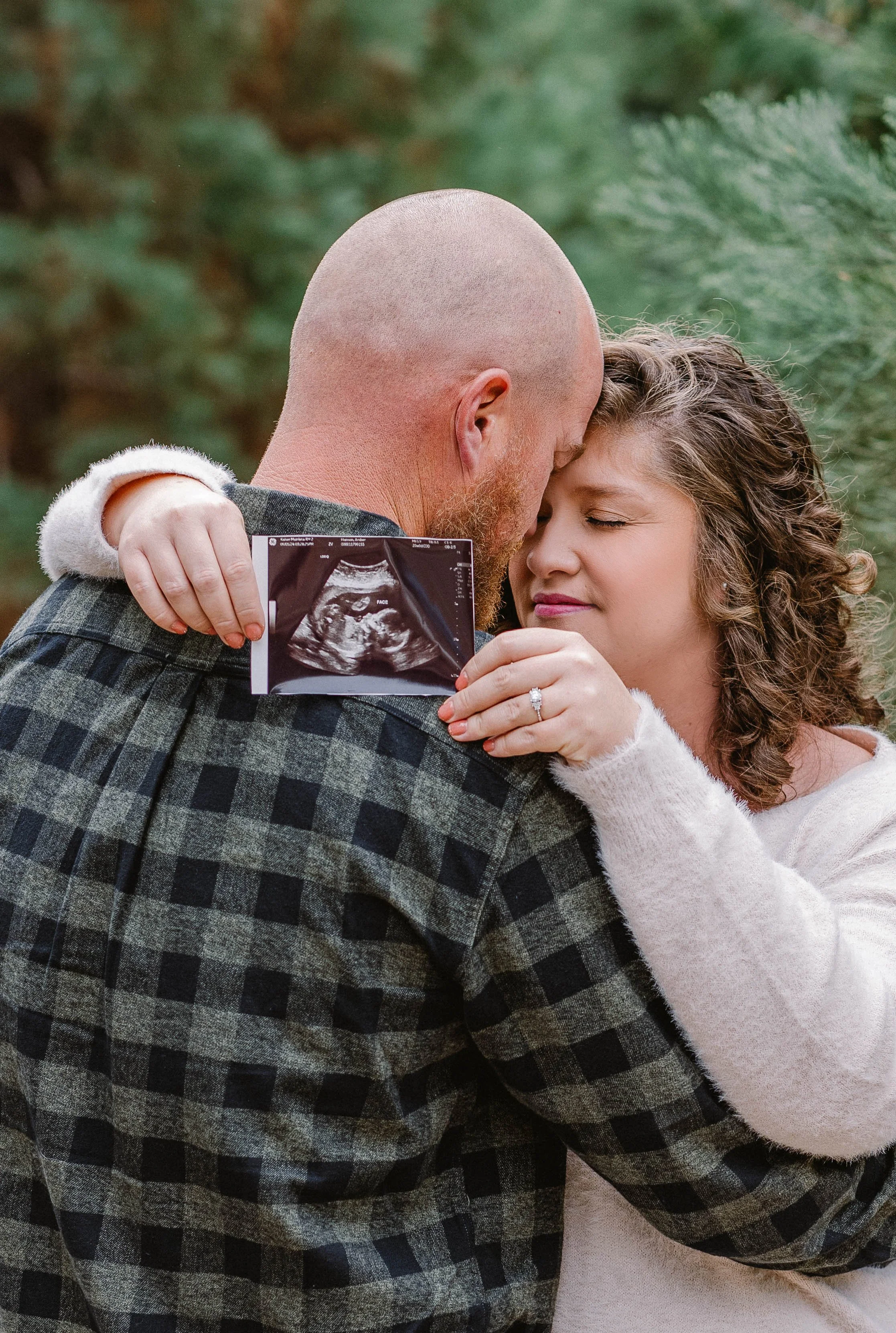 A couple embraces outdoors, holding an ultrasound image, with a blurred green background.