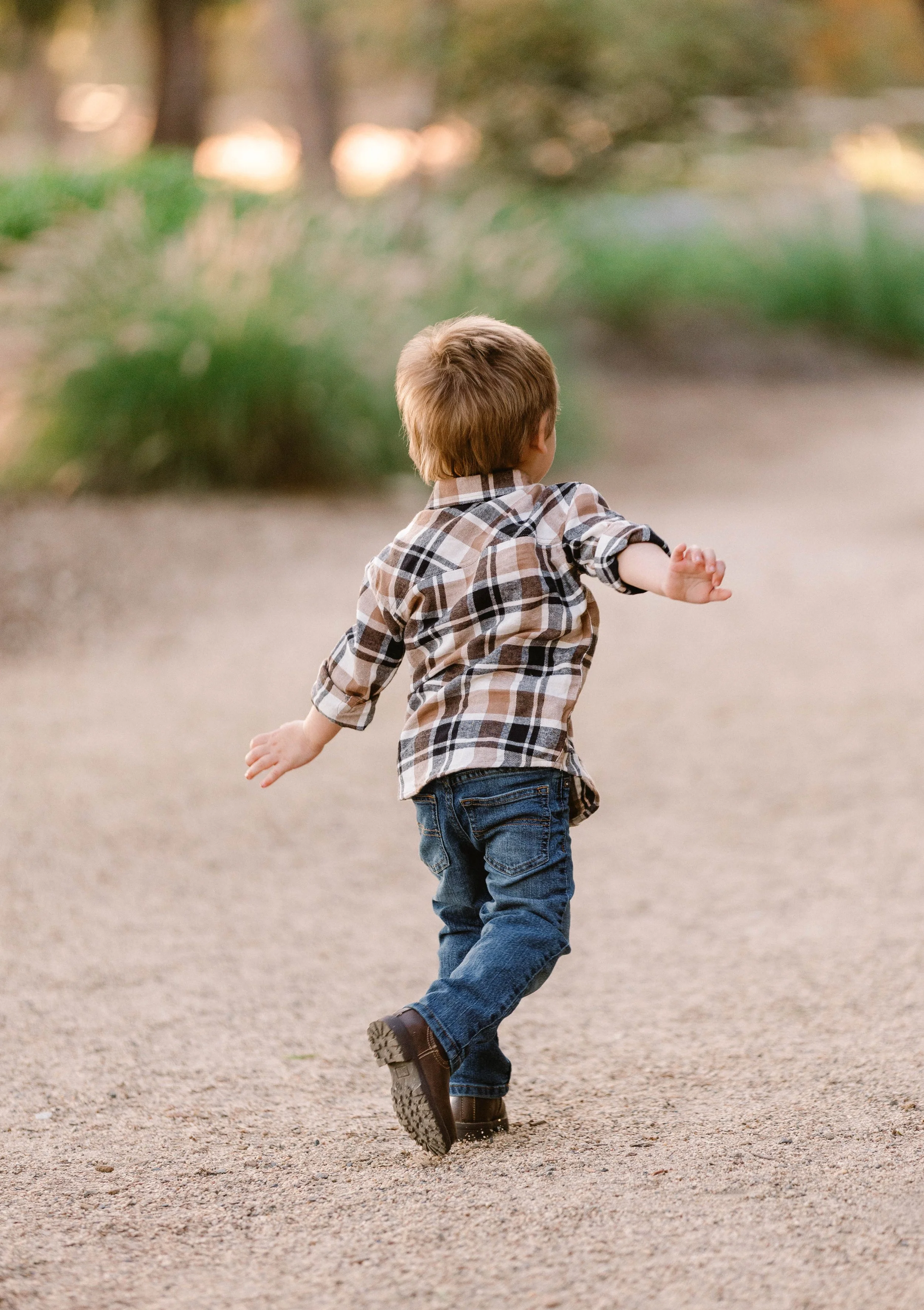 A young boy running on a dirt path outdoors, wearing a plaid shirt, jeans, and boots, with arms extended.