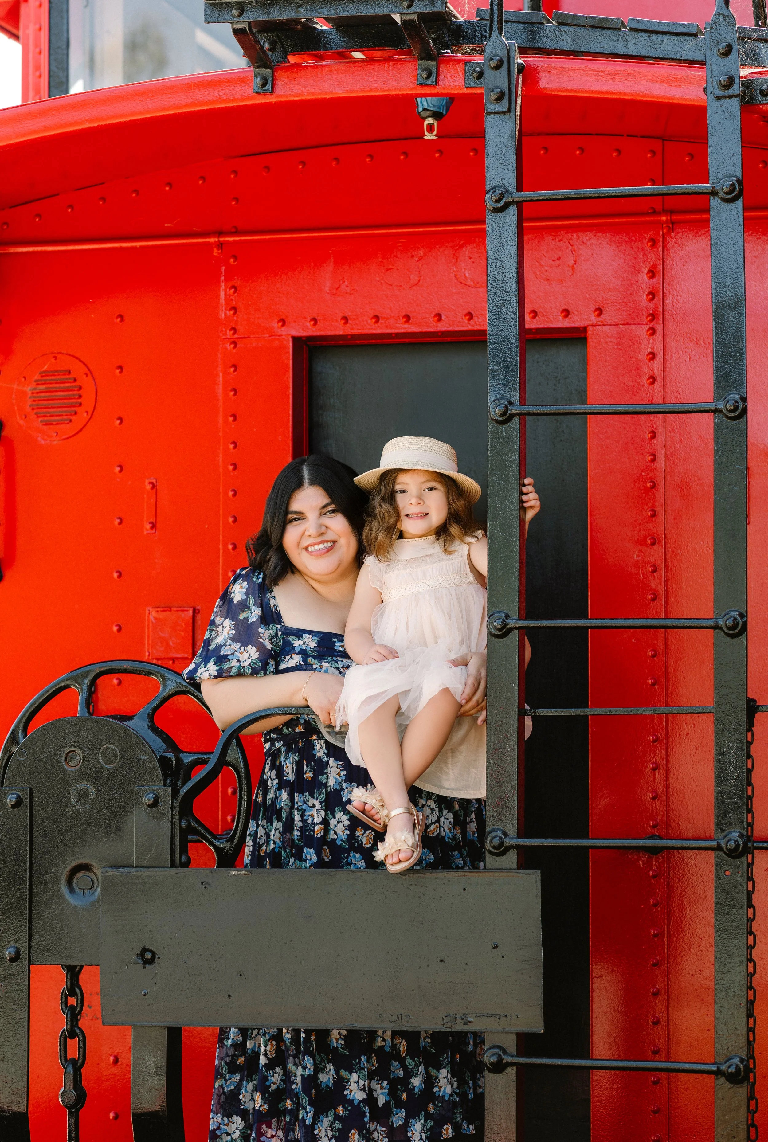 A woman and a young girl sitting on a train's platform, with the woman smiling and the girl wearing a sunhat and light-colored dress, against a bright red train car.