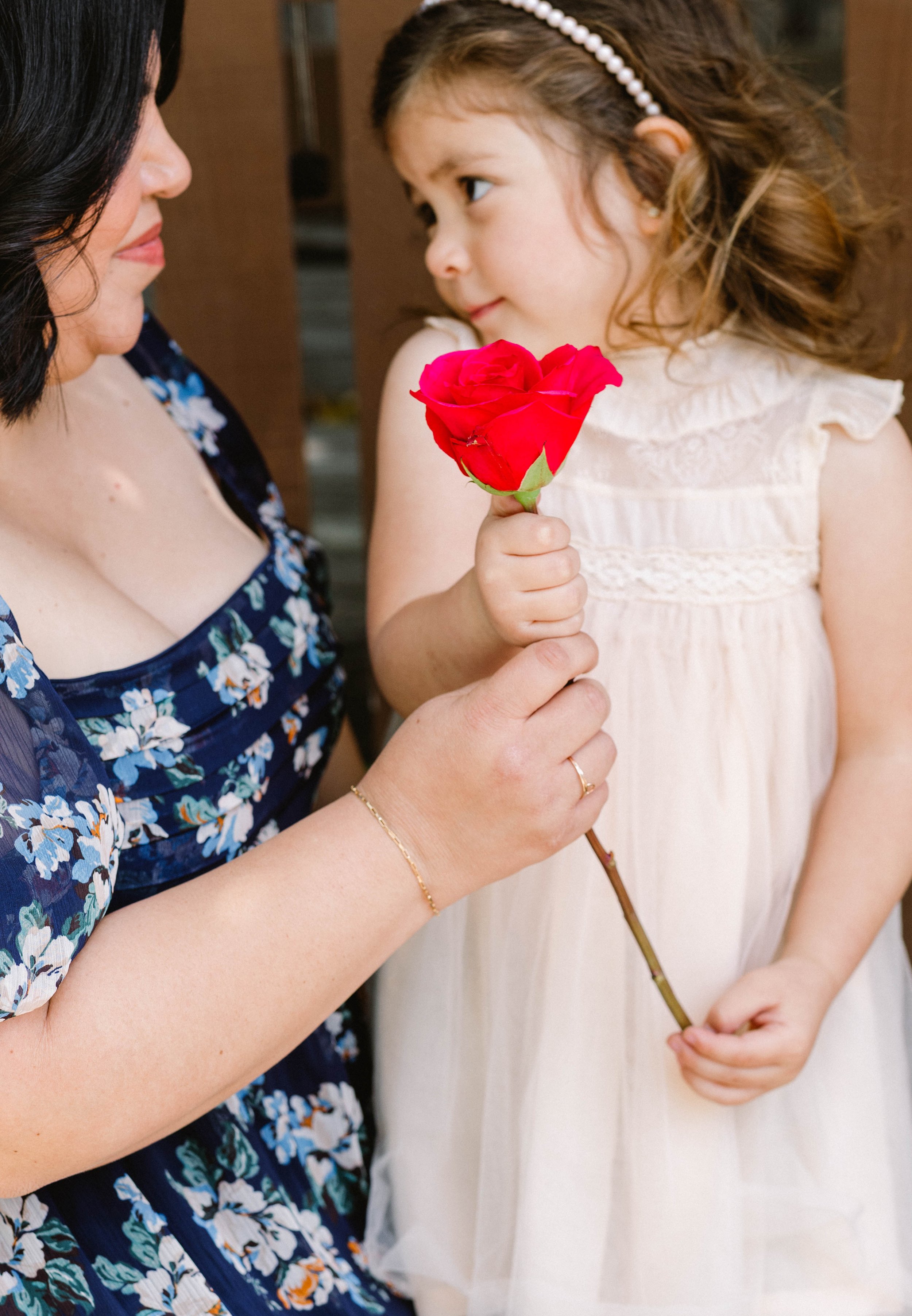 A woman in a blue floral dress holding a red rose while a young girl in a white dress looks at her and holds the rose