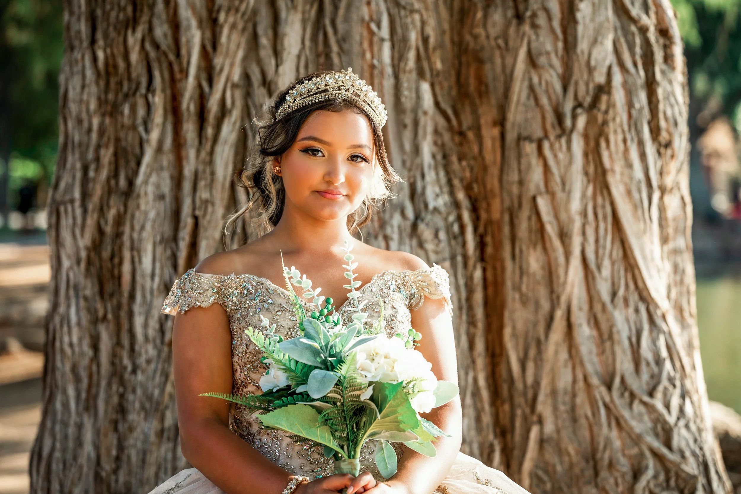 A young woman in a wedding dress and tiara holding a bouquet of flowers outdoors beside a large tree trunk.