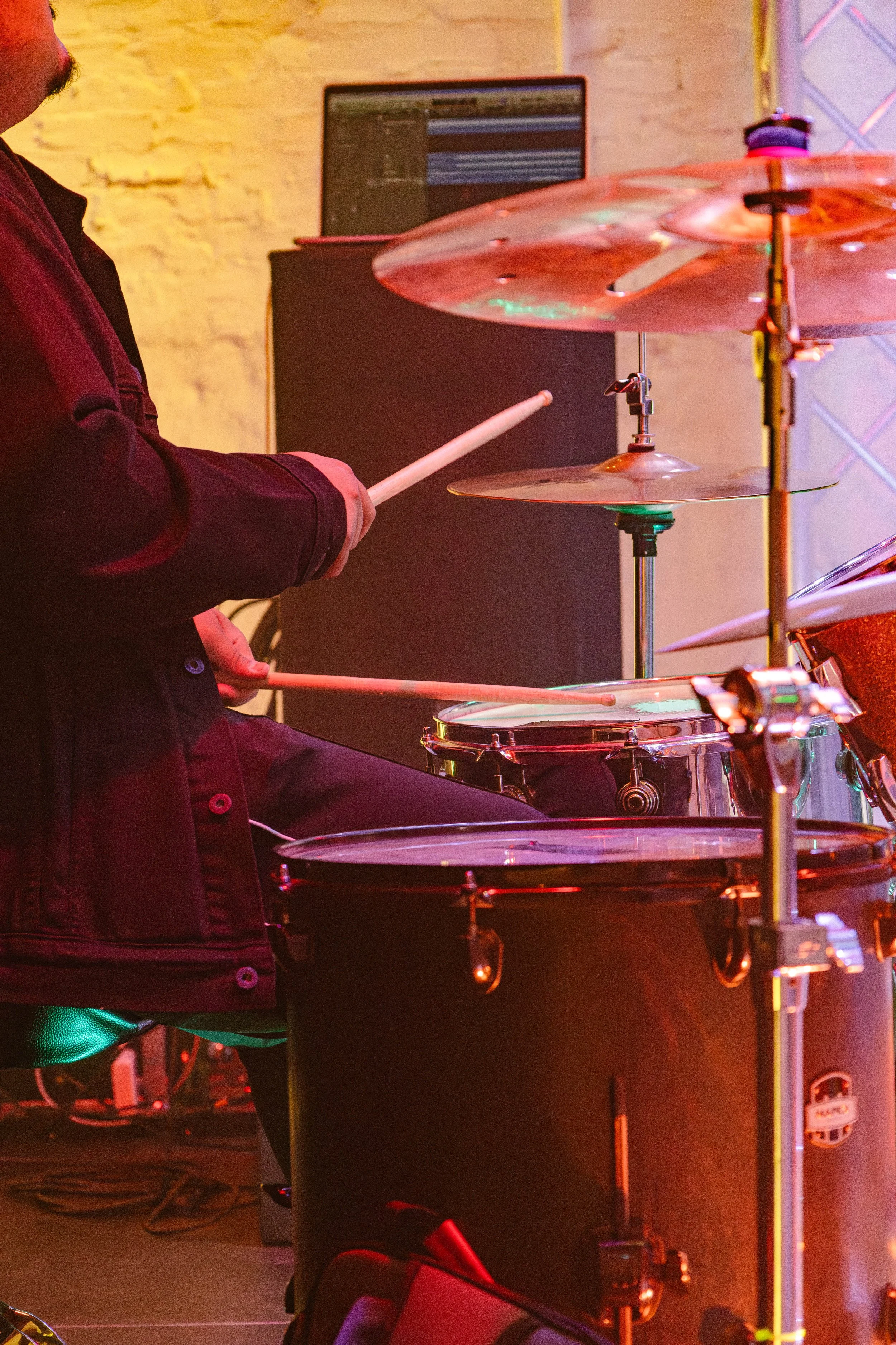 Close-up of a person playing drums, with a drum set and a music production computer in the background, illuminated by stage lighting.