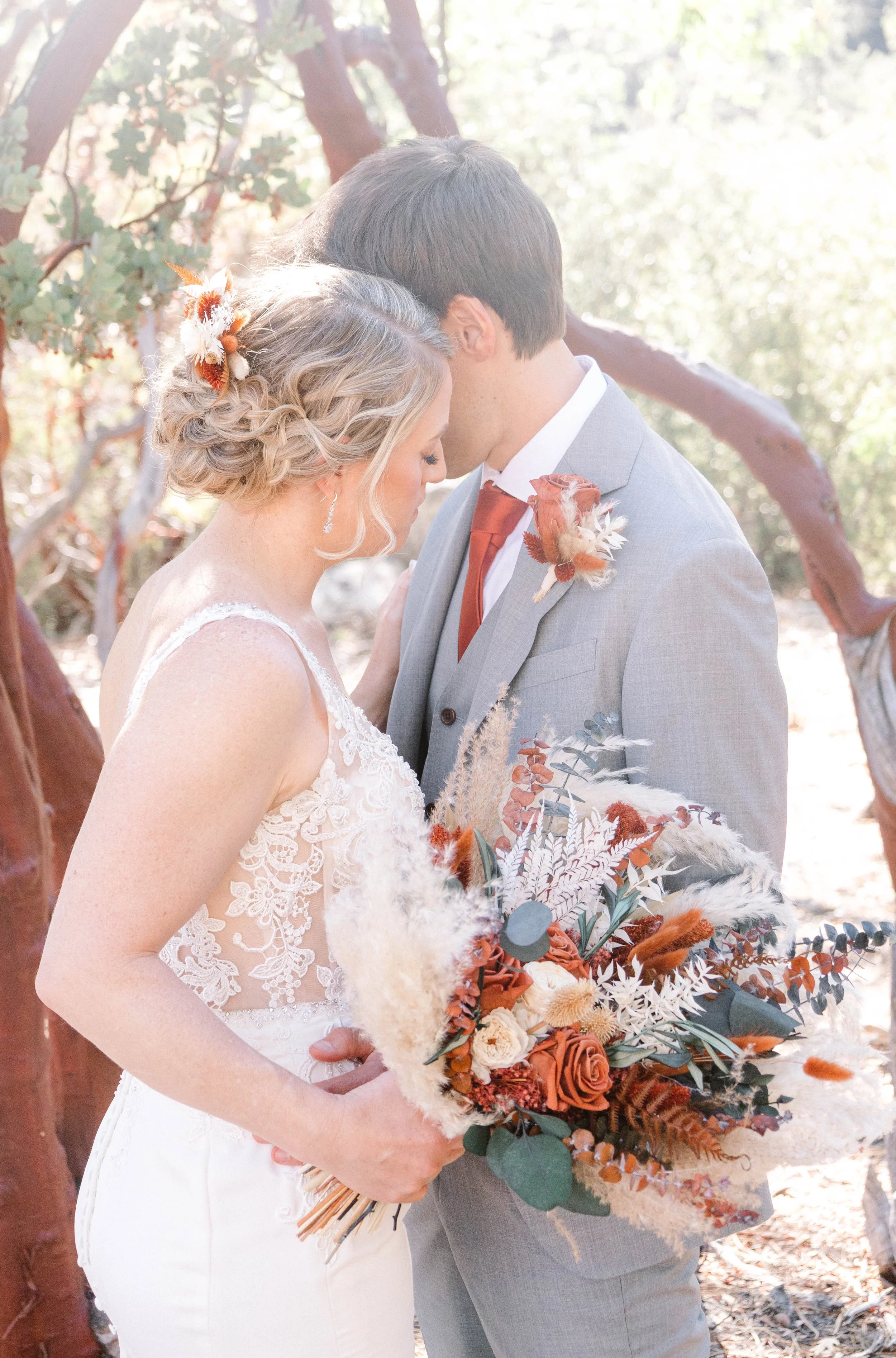 A bride and groom sharing a tender moment outdoors, with their foreheads touching and eyes closed. The bride holds a large bouquet of dried and fresh flowers in shades of orange, cream, and green. The couple is dressed in wedding attire, and the setting features sunlight filtering through trees.