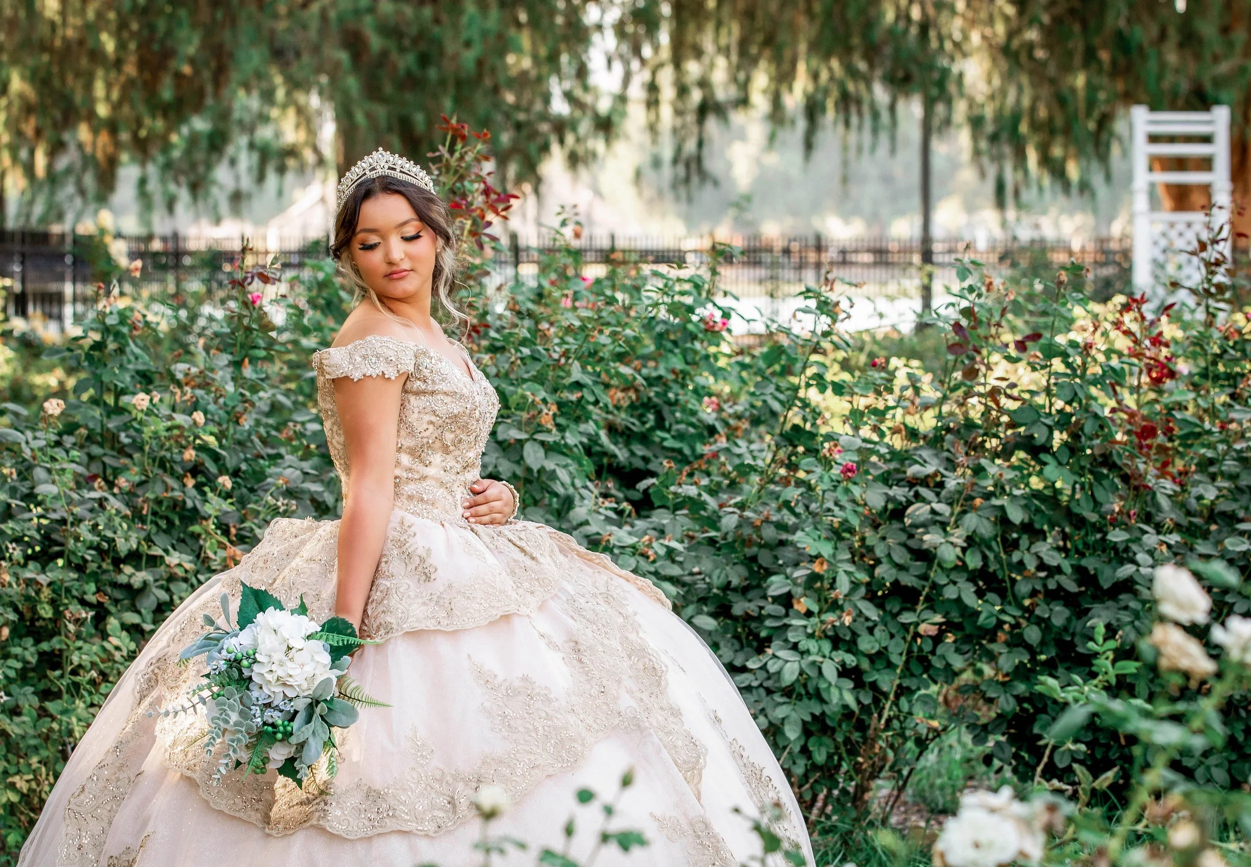 A young woman in an elegant beige and gold fancy dress with lace details and a tiara, holding a bouquet of white and green flowers, standing in a garden with green bushes and blooming roses.