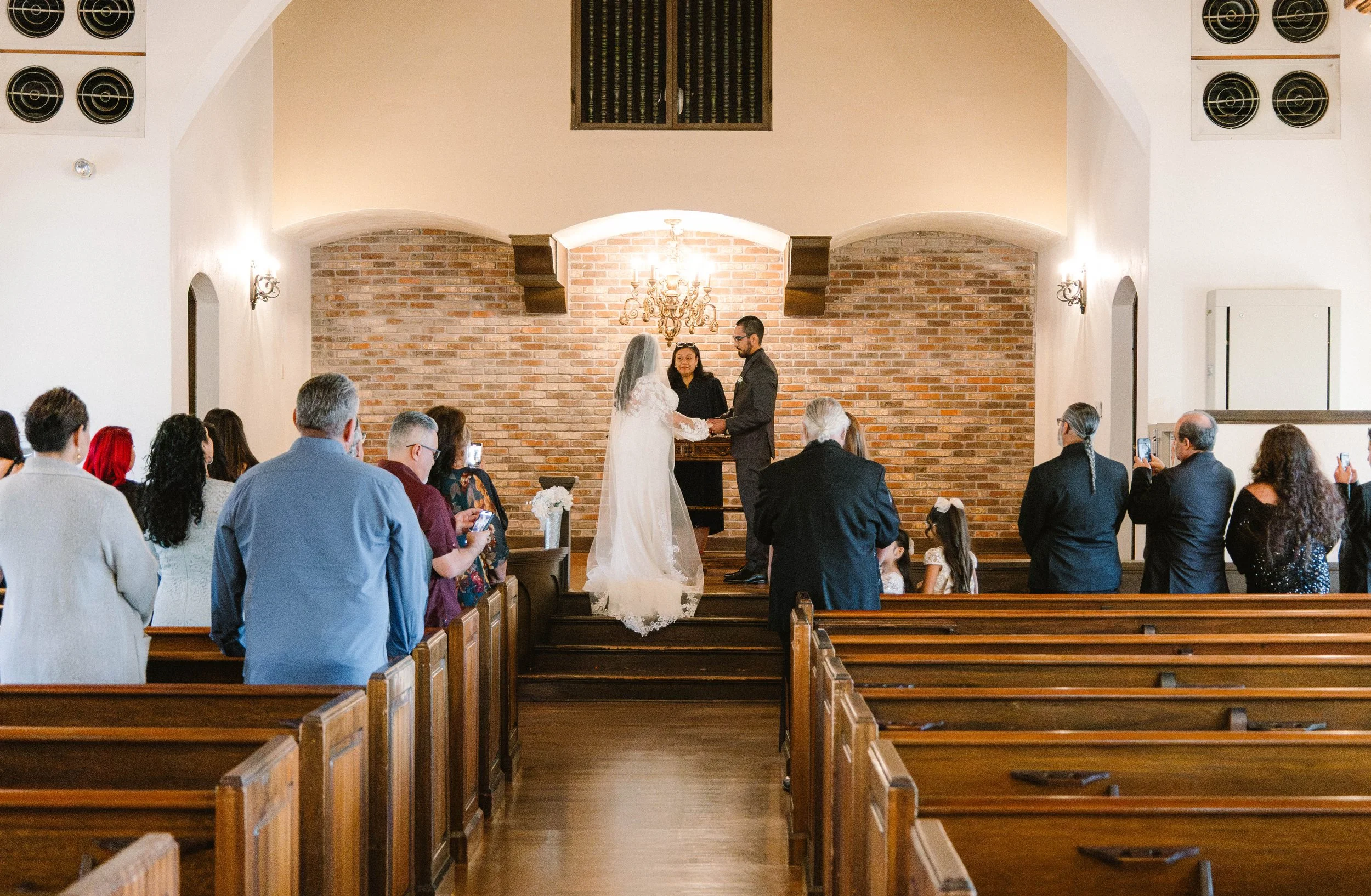 A wedding ceremony taking place in a church with the bride and groom standing at the altar and exchanging vows, surrounded by friends and family seated in wooden pews.