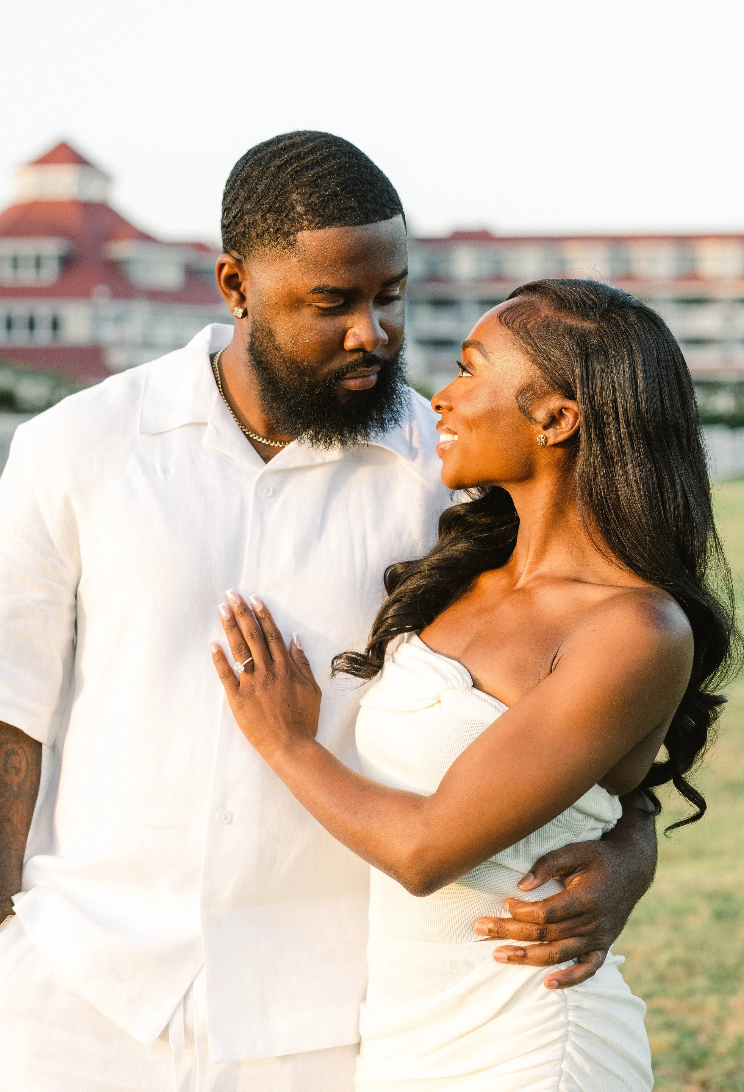 A couple standing close together outdoors during sunset, gazing into each other's eyes, with the woman wearing a white strapless dress and the man in a white shirt.