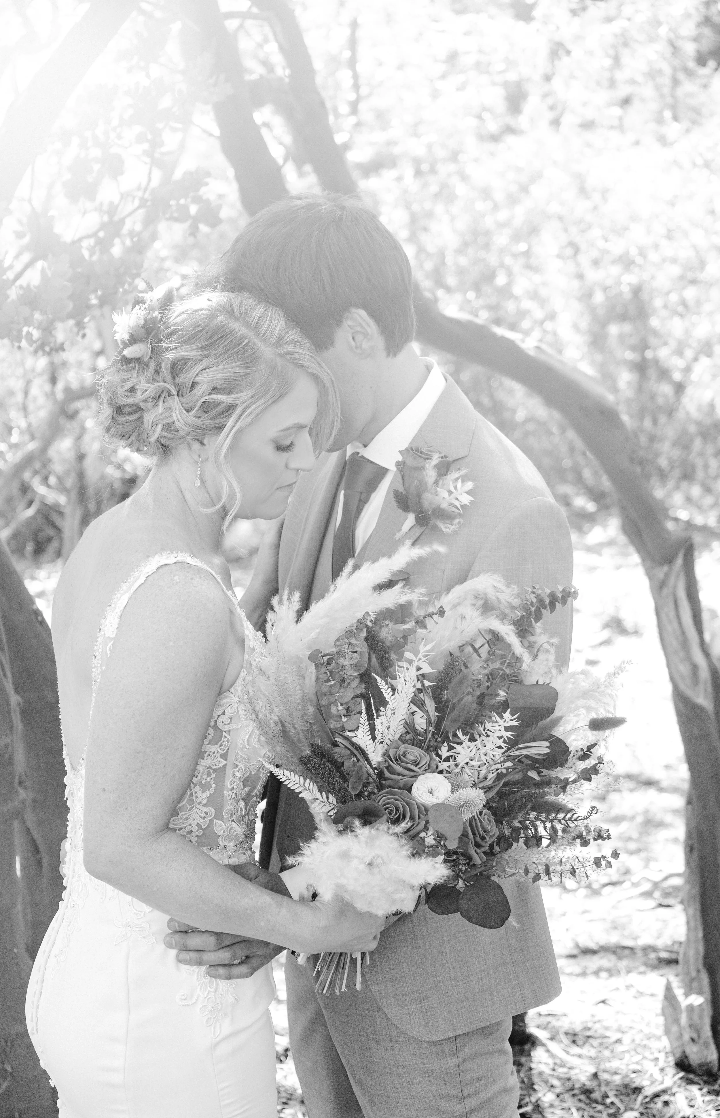 A black-and-white photo of a bride and groom standing close, with heads bowed and eyes closed, holding a bouquet of flowers outdoors surrounded by trees.