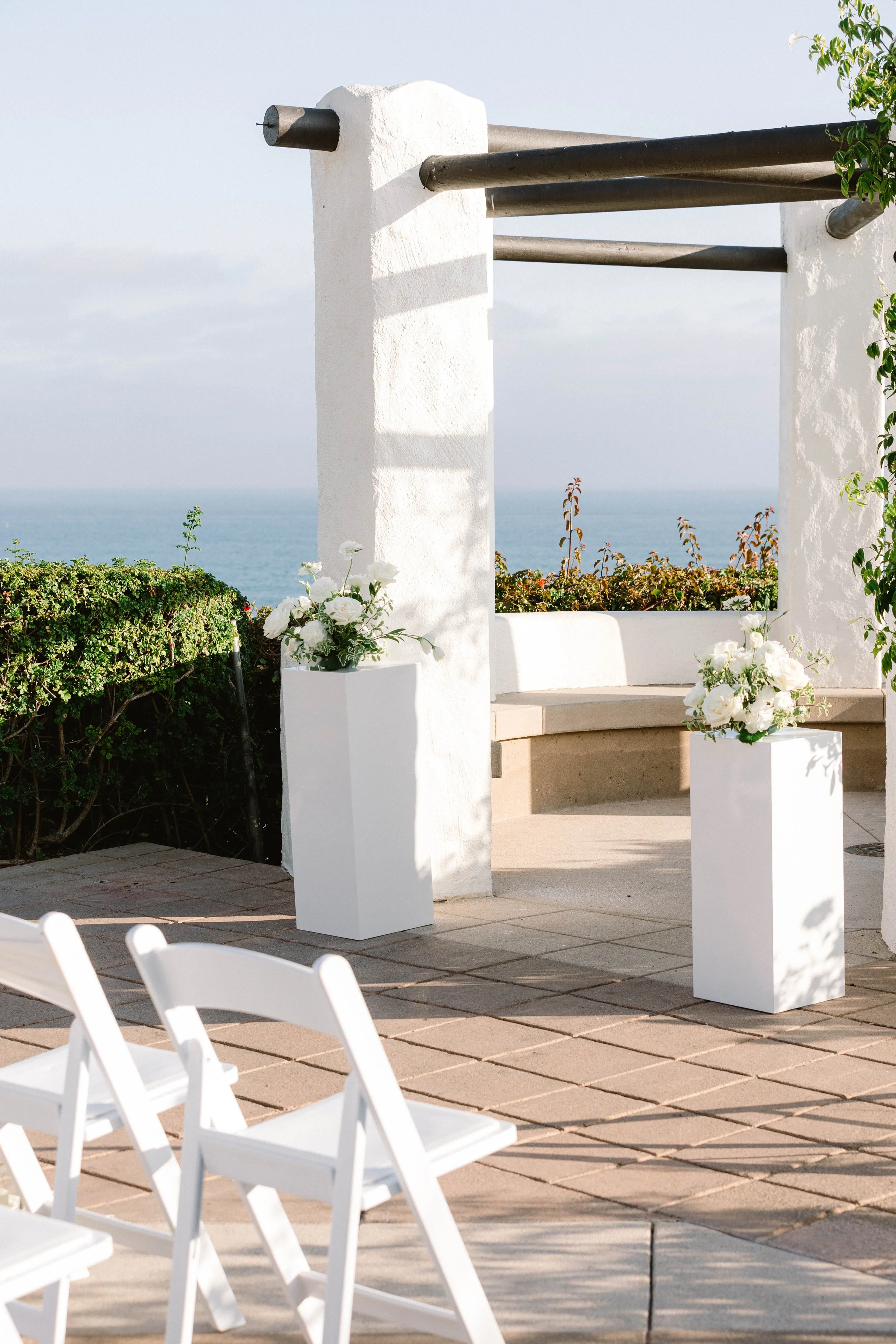 Decorative outdoor ceremony setup with white chairs, white flower arrangements on tall pedestals, and a white arch structure overlooking the ocean.