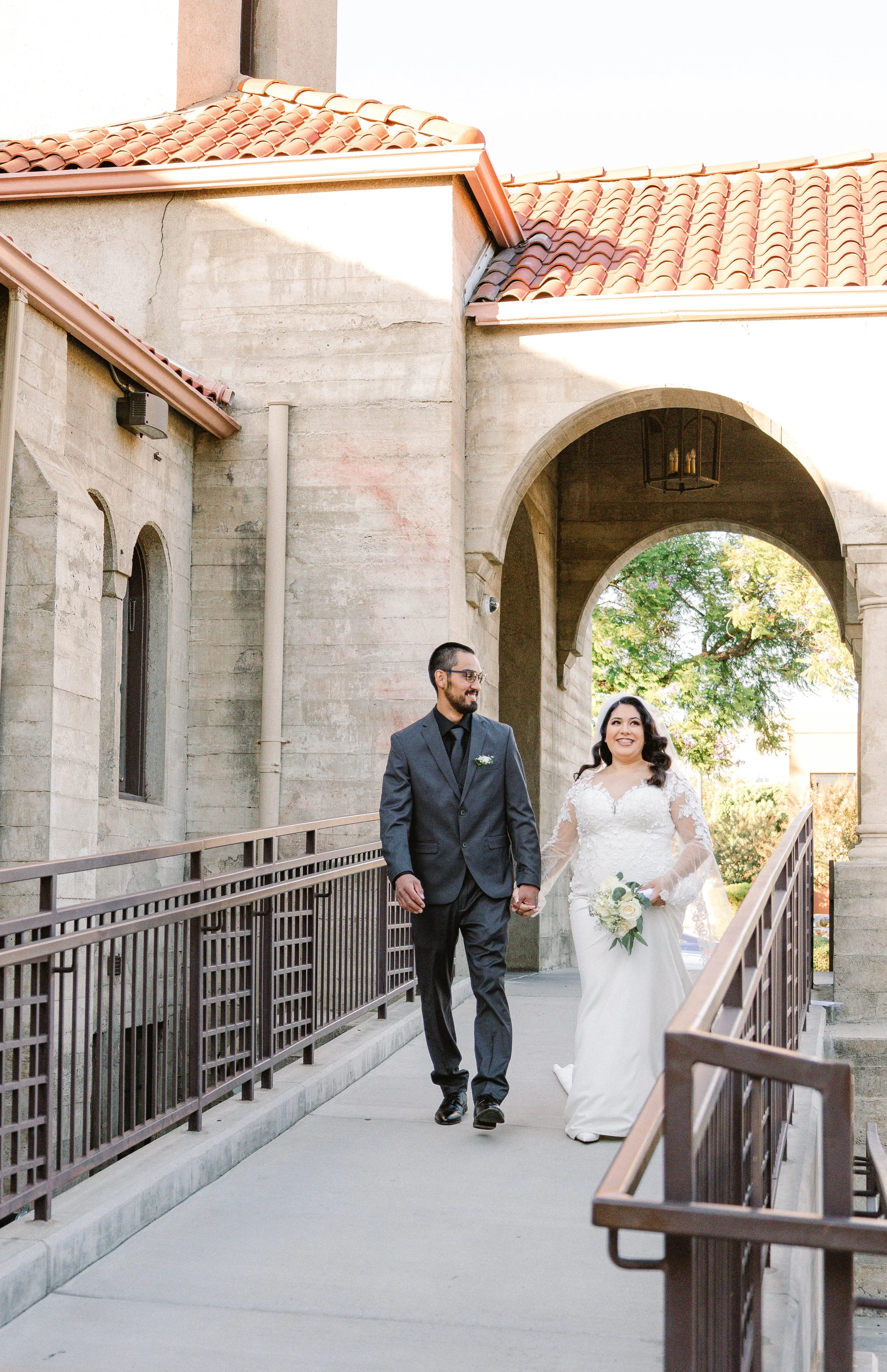 A bride and groom walking hand in hand outside a stone building with an arched entryway and a tree in the background.