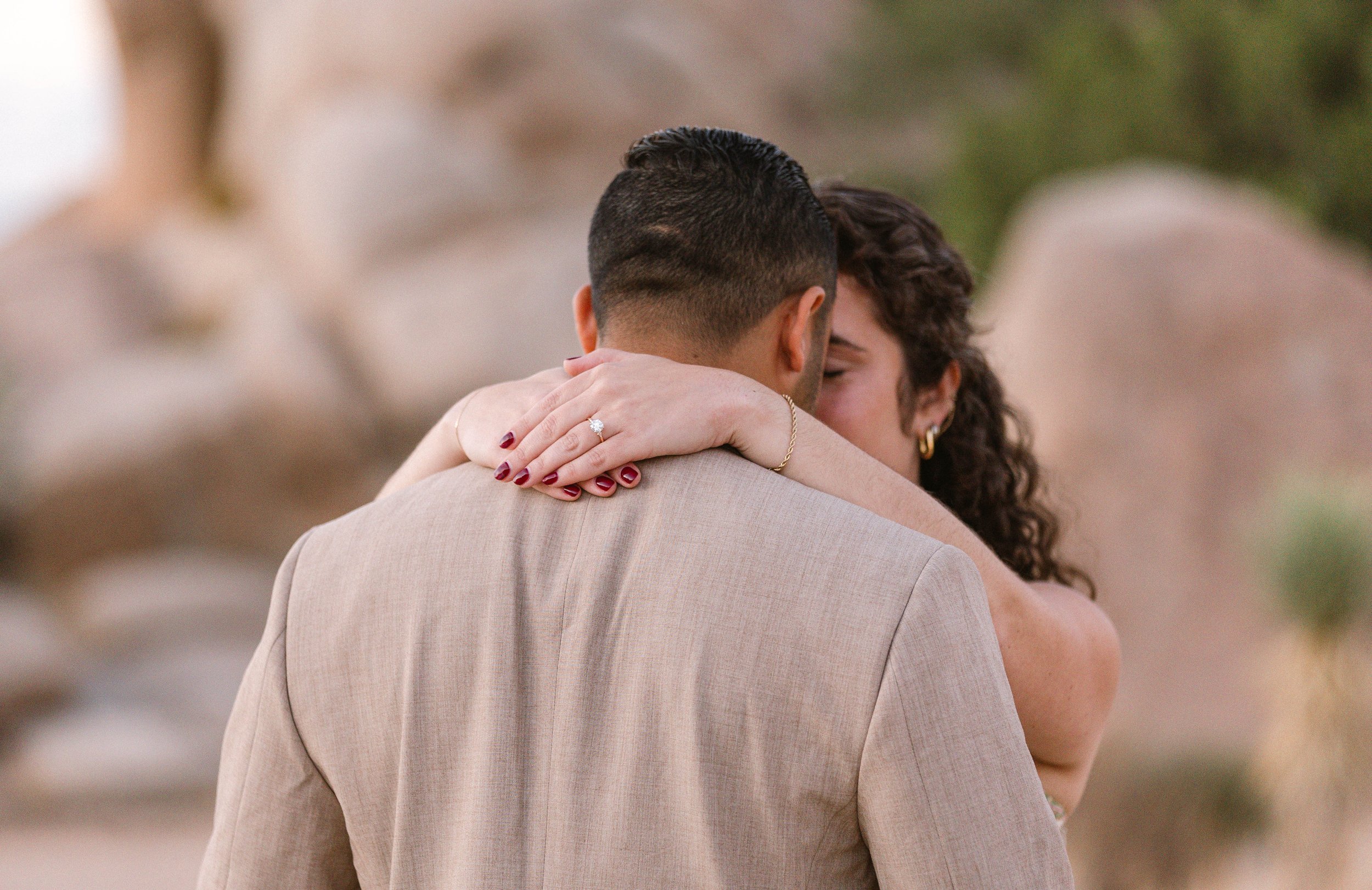 A couple hugs, with the woman showing off her engagement ring in the foreground