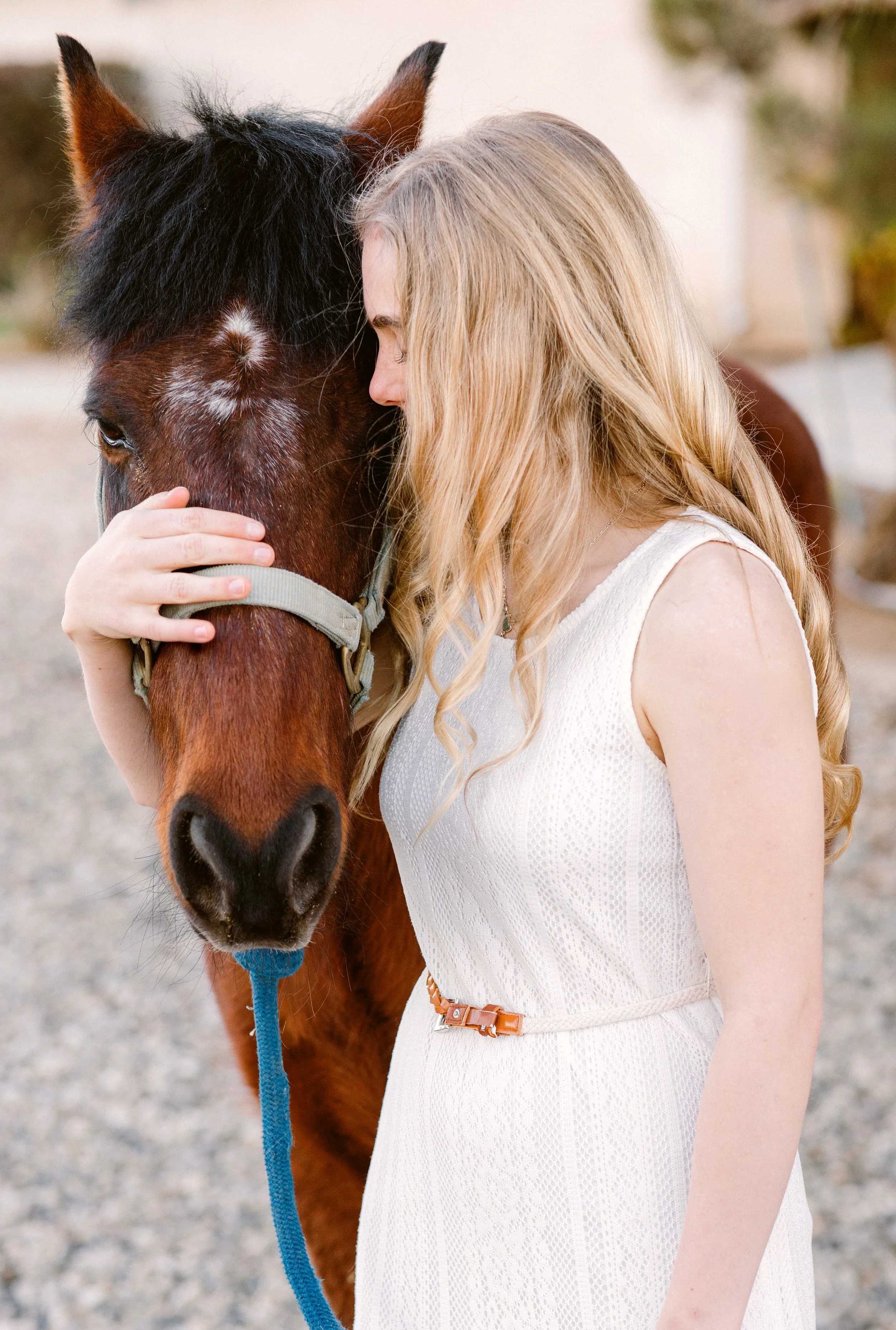 A woman with long blonde hair in a white sleeveless dress gently touching and leaning her forehead against a brown horse with a black mane.