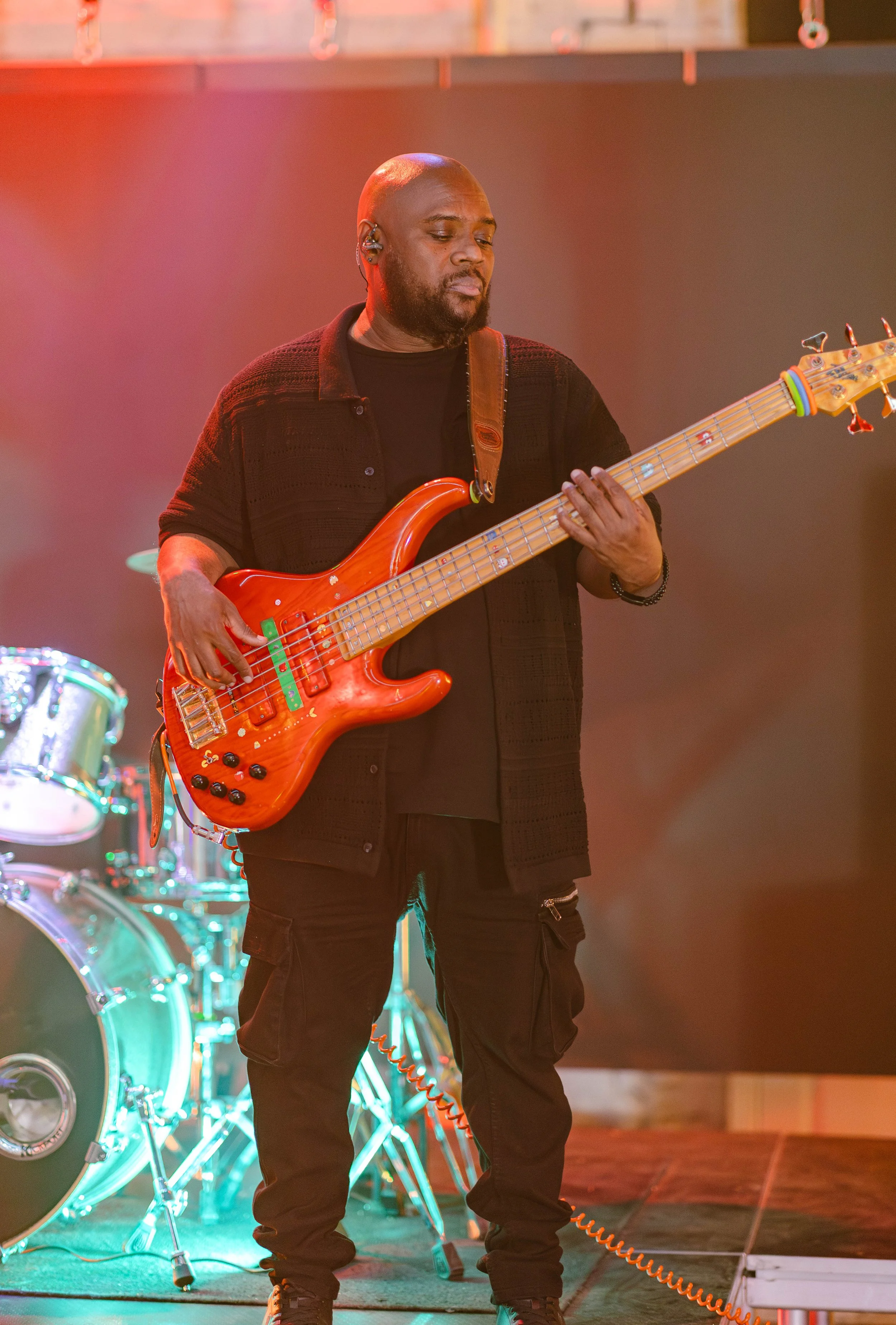 A man playing a red electric bass guitar on stage with drums in the background.