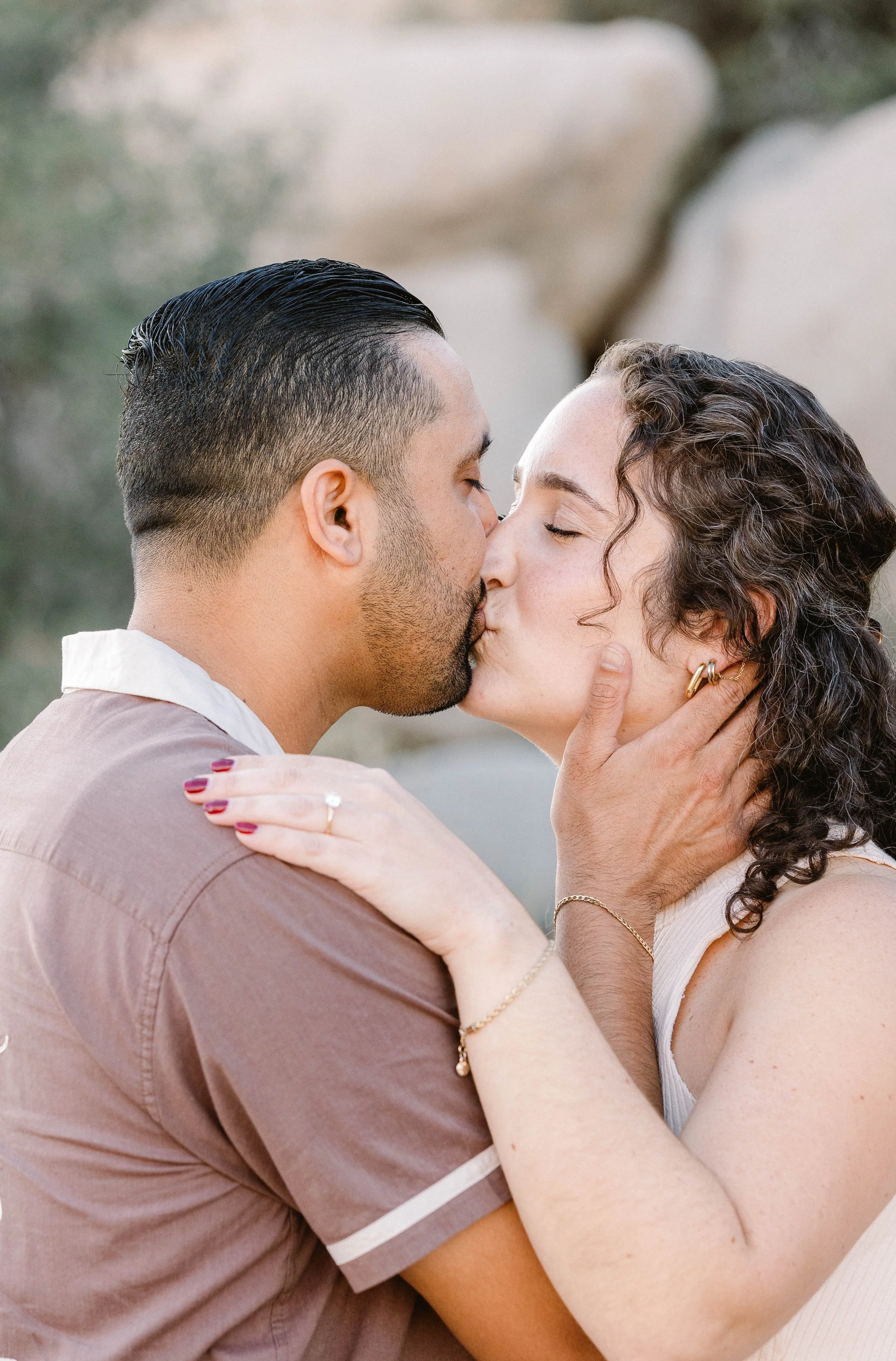 A couple kissing outdoors, with the man holding the woman's face gently and the woman holding onto his shoulder, both appearing happy and affectionate.