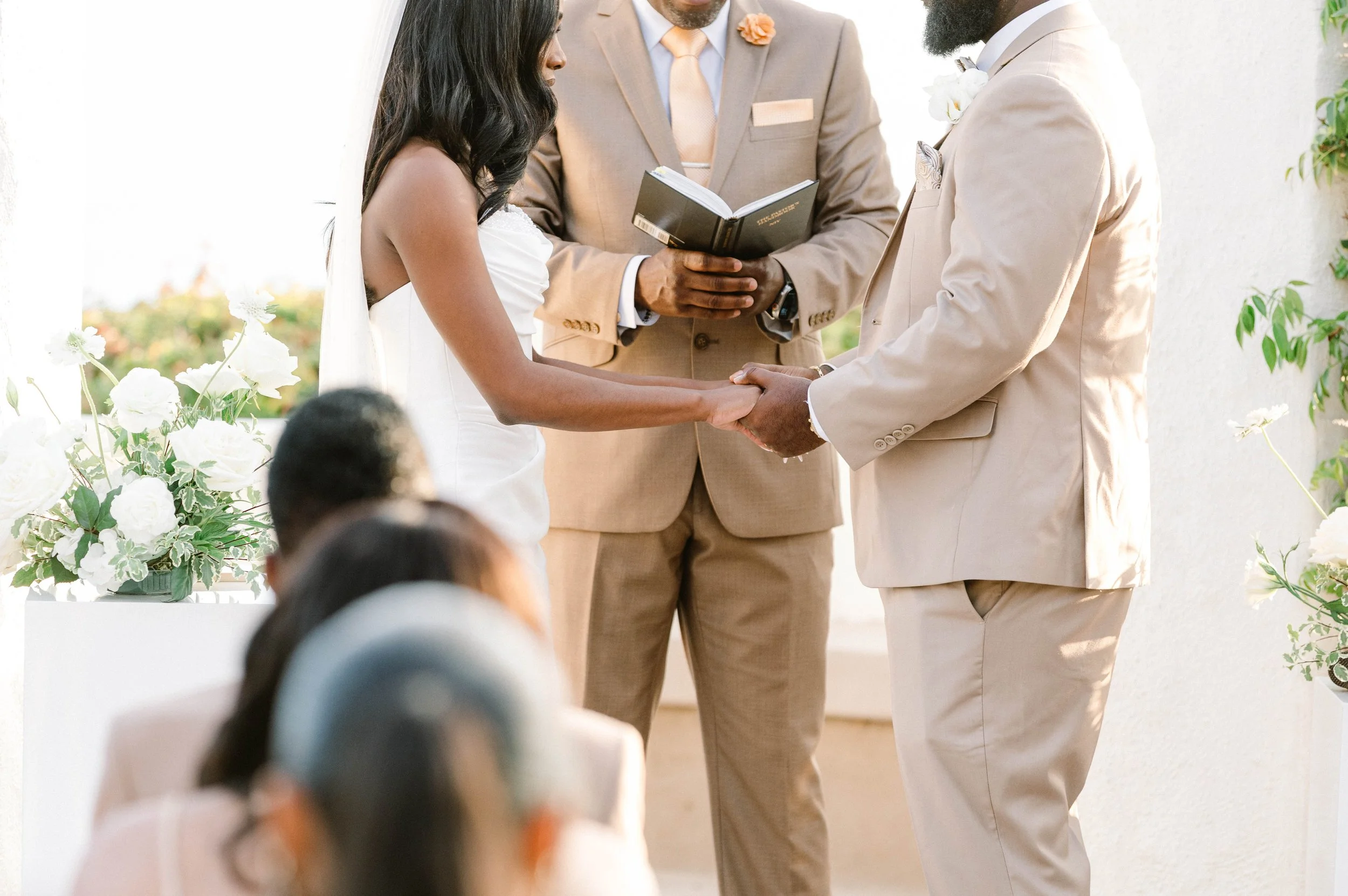 A couple getting married holding hands during their wedding ceremony, with an officiant standing behind them, holding a book, in an outdoor setting decorated with white flowers.