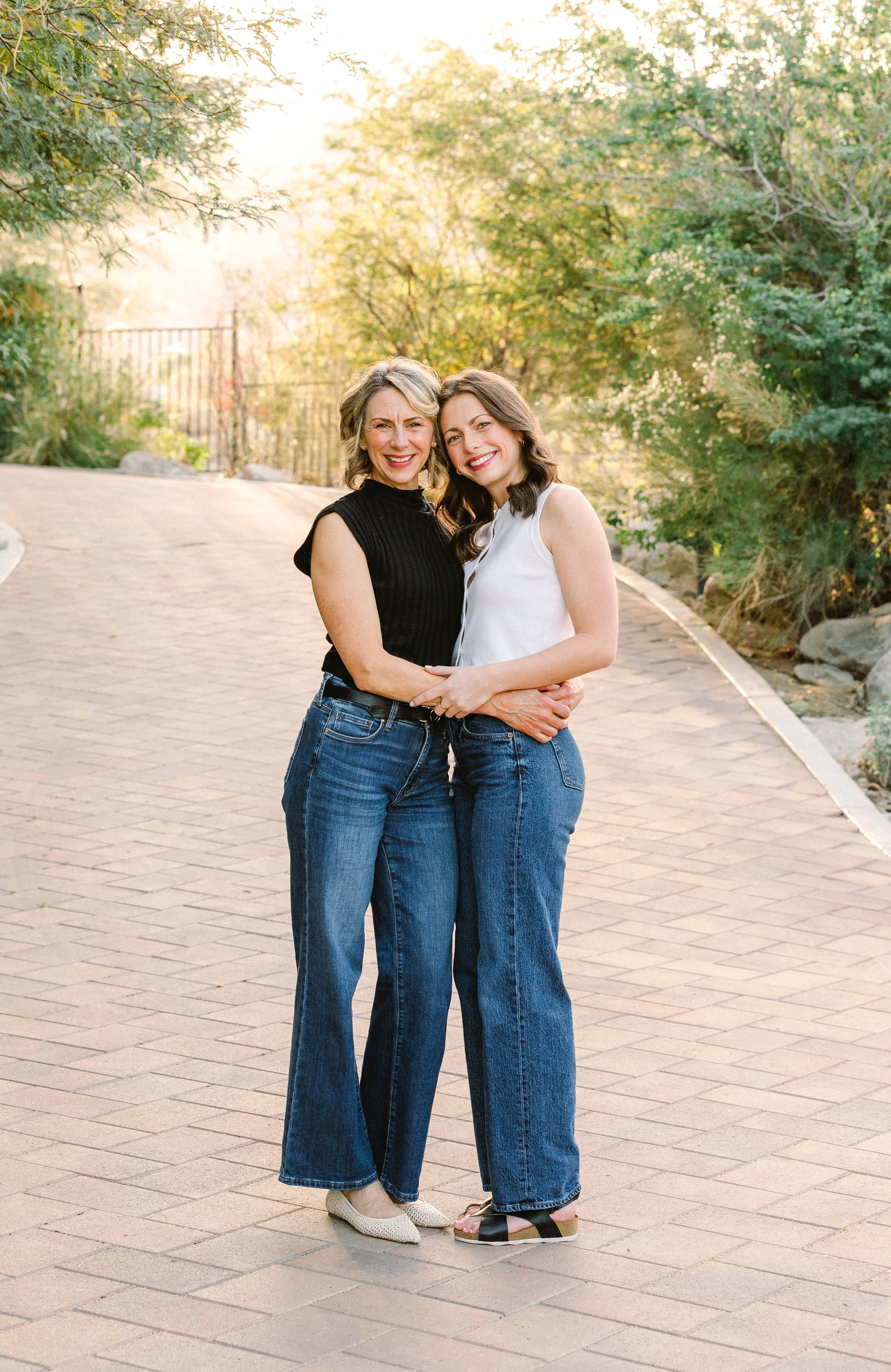 Two women standing close together on a brick pathway, smiling, with greenery and trees in the background during a sunny day.