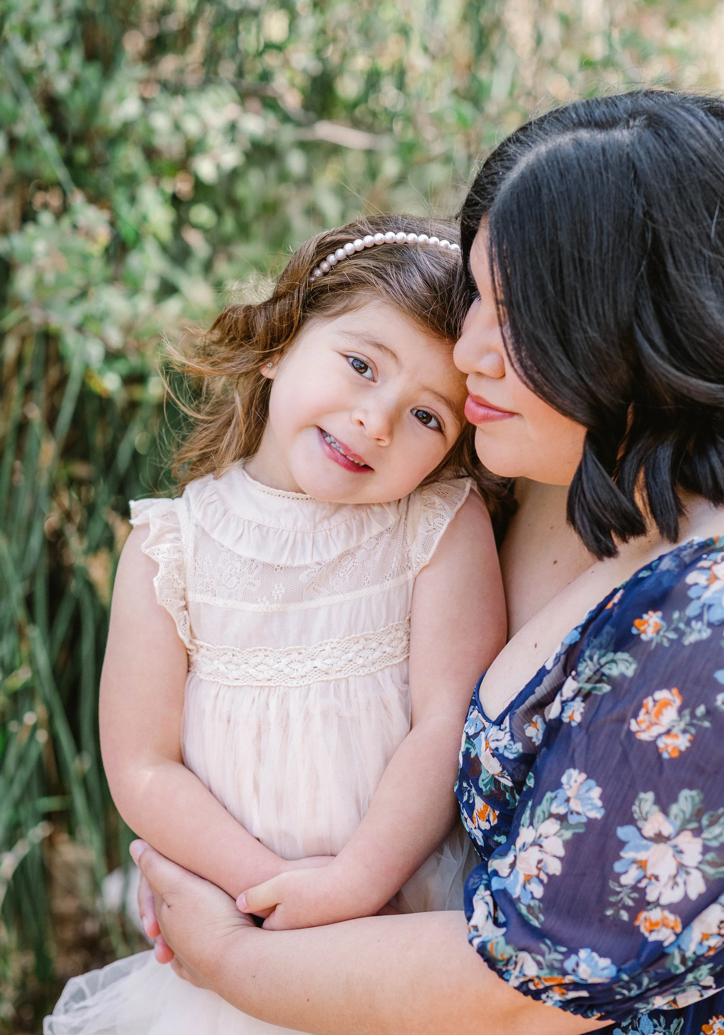 A young girl with brown hair and a pearl headband is in an outdoor setting, holding hands with a woman with dark hair in a blue floral dress, both with gentle expressions.