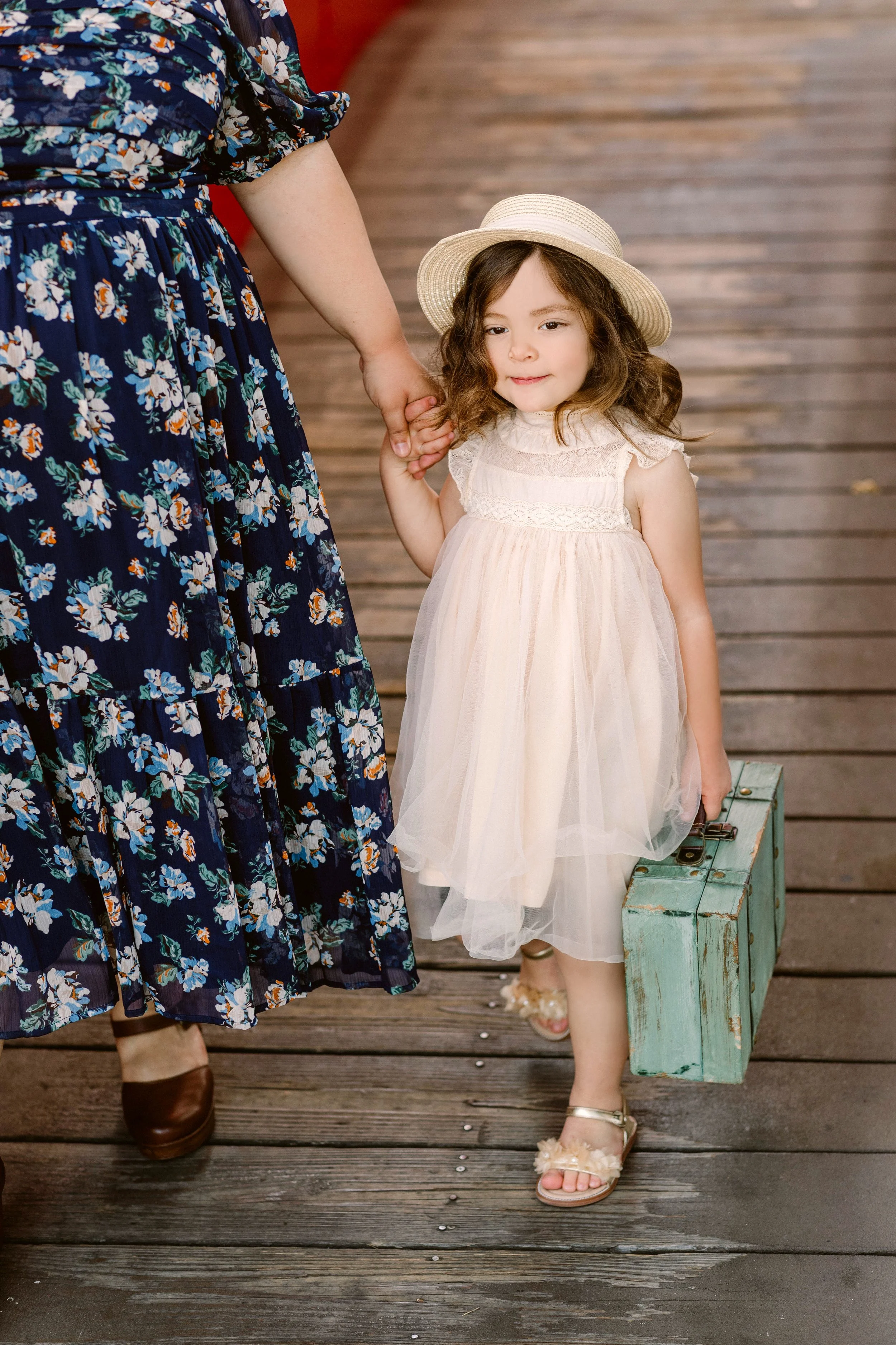 Young girl in a white dress and sunhat holding a teal wooden suitcase, walking hand-in-hand with an adult on a wooden boardwalk.