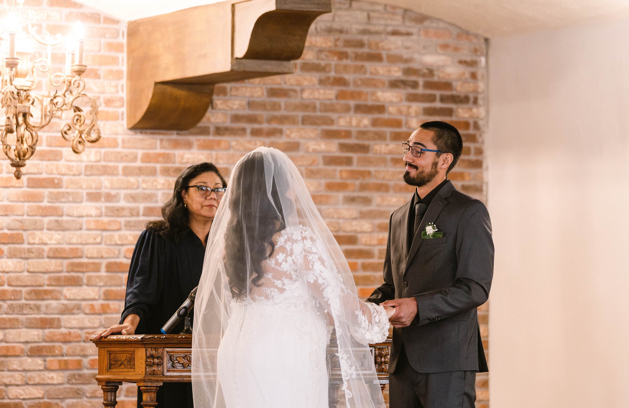 A bride and groom holding hands during their wedding ceremony, with a woman officiant standing behind a wooden podium, in a room with a brick wall and a decorative wall sconce.