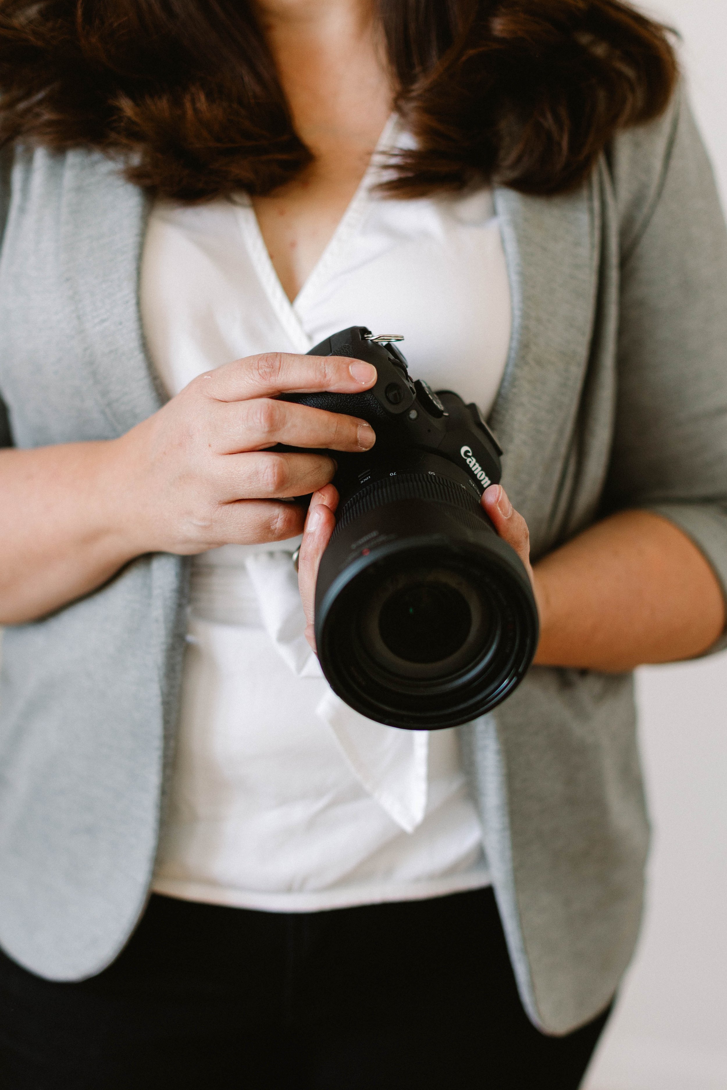Person holding a Canon DSLR camera, dressed in a white top and gray blazer.