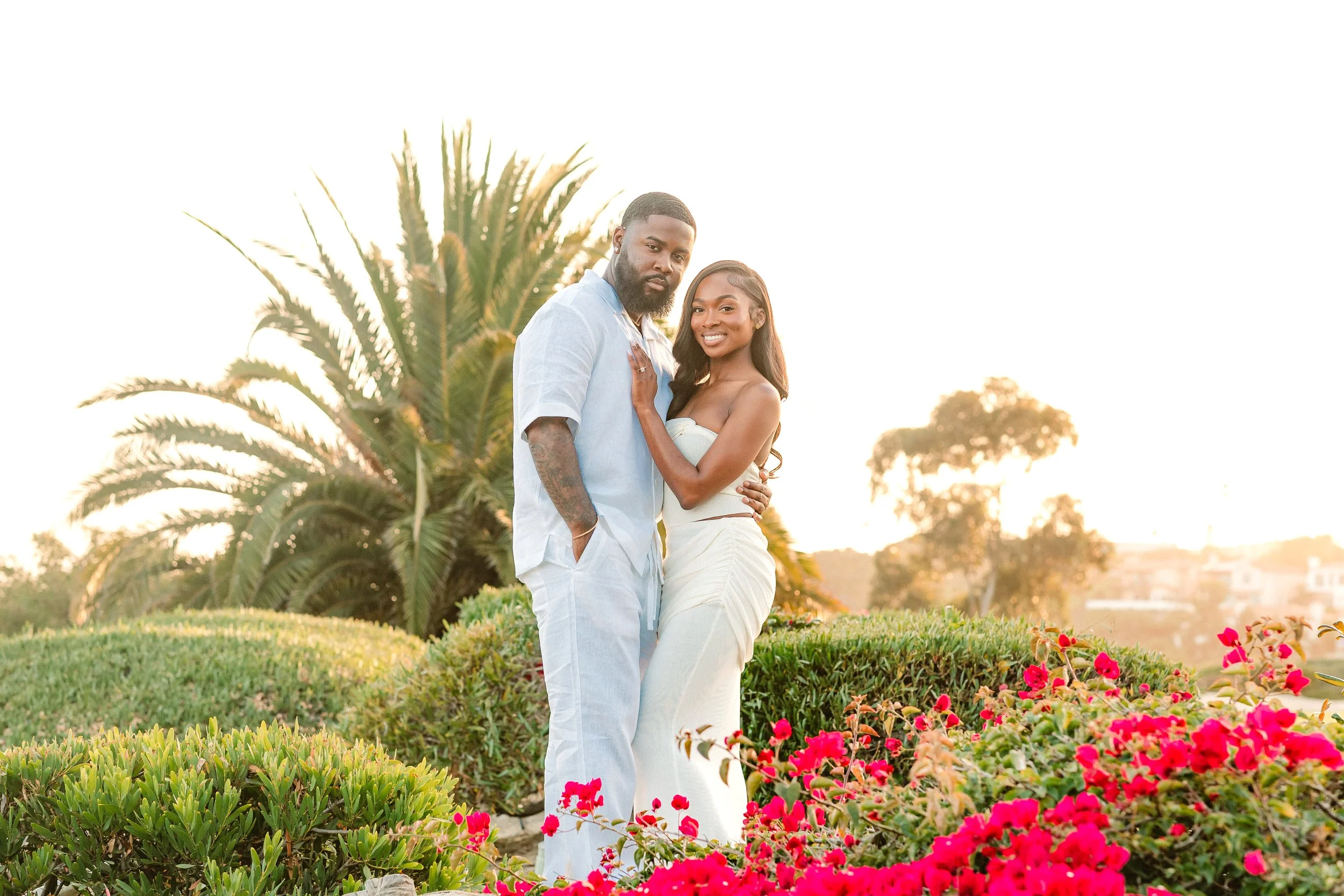 A smiling couple standing close together outdoors during sunset, surrounded by vibrant pink flowers and green bushes.