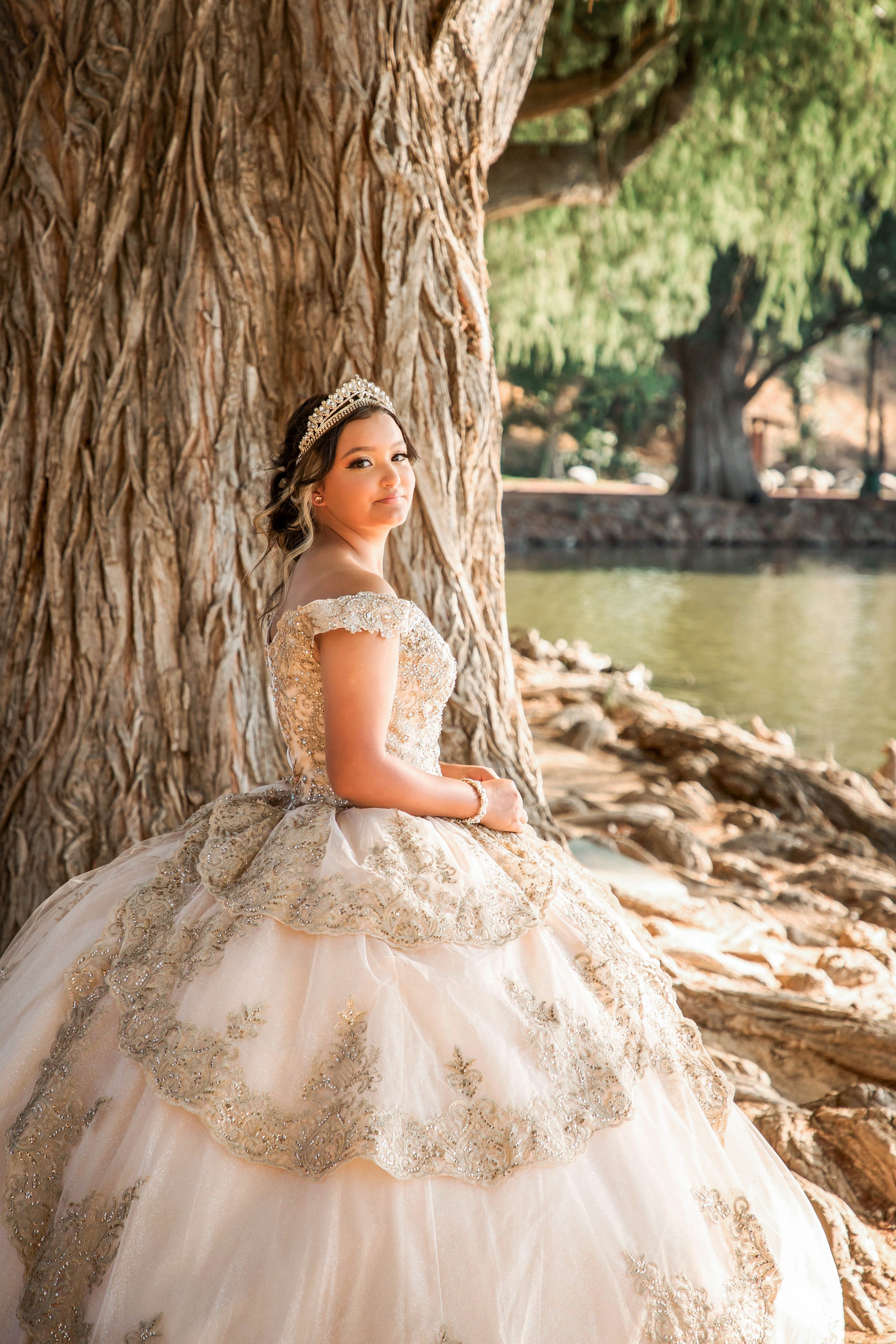 A young woman in an ornate, cream-colored gown with gold embroidery, sitting against a large tree near a pond, wearing a tiara and jewelry.