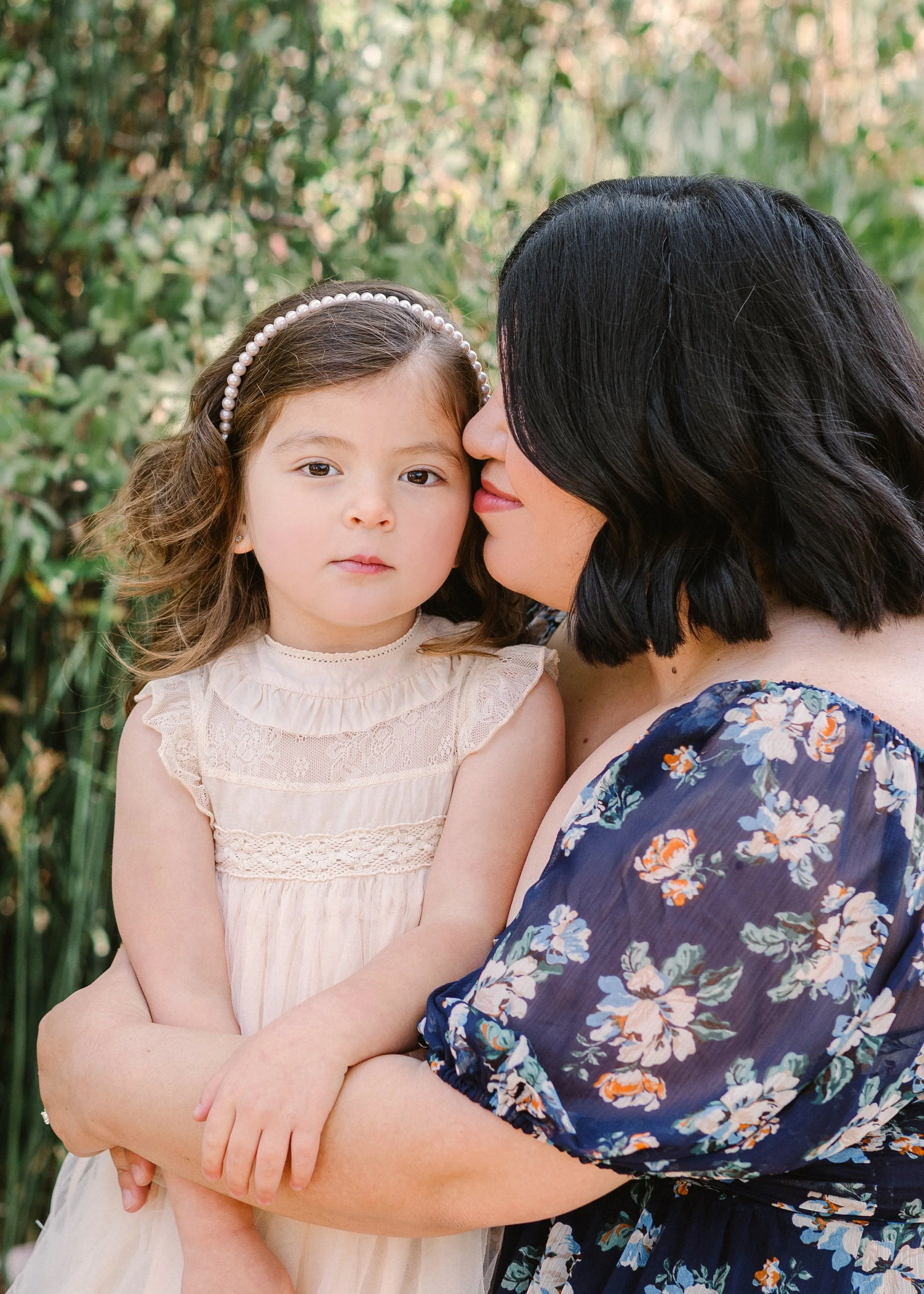 A woman with dark hair and a floral dress holding a young girl with a pearl headband, in an outdoor setting with greenery.