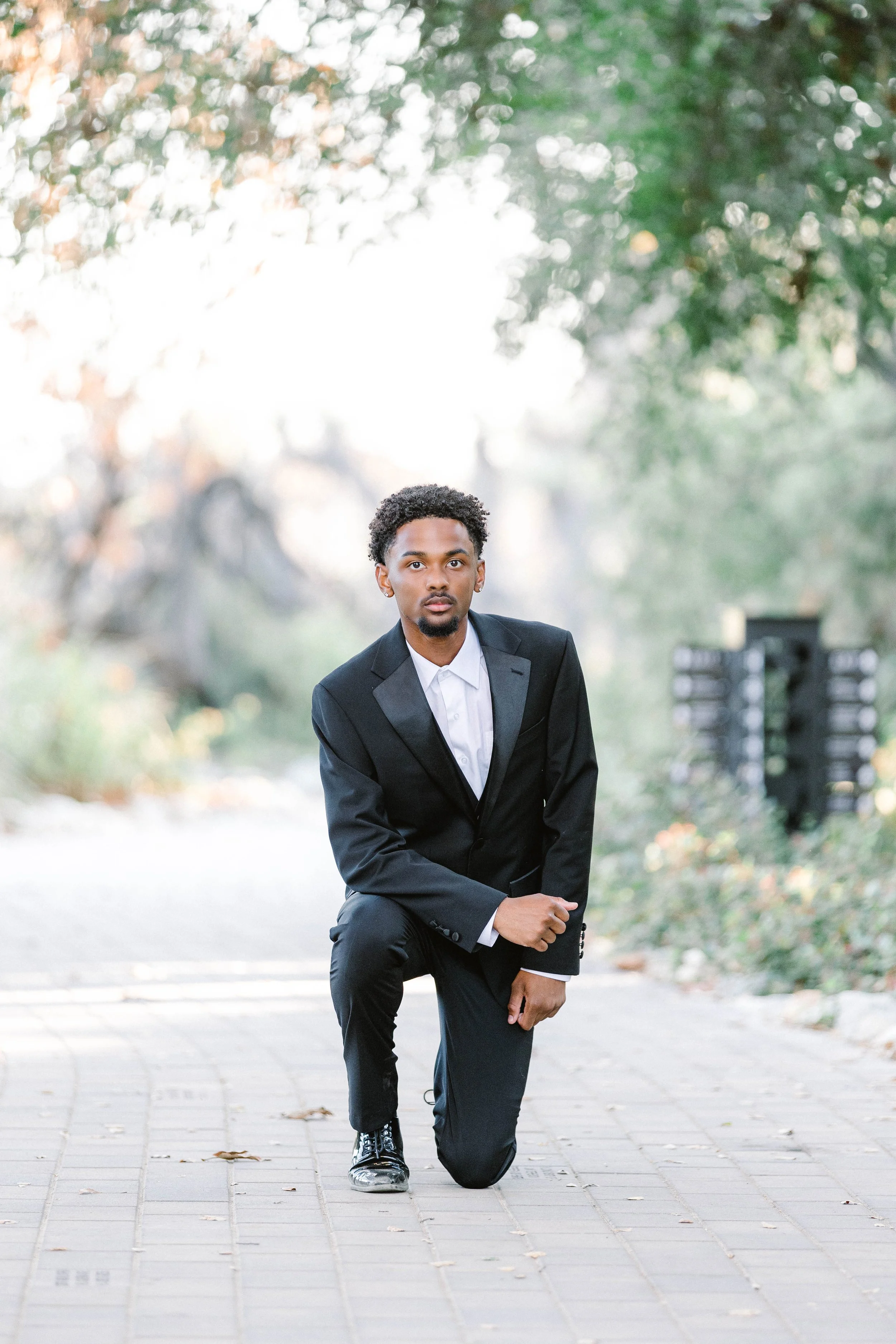 A young man dressed in a black suit and white shirt kneeling on one knee on a paved path in a park-like setting with trees and greenery in the background.