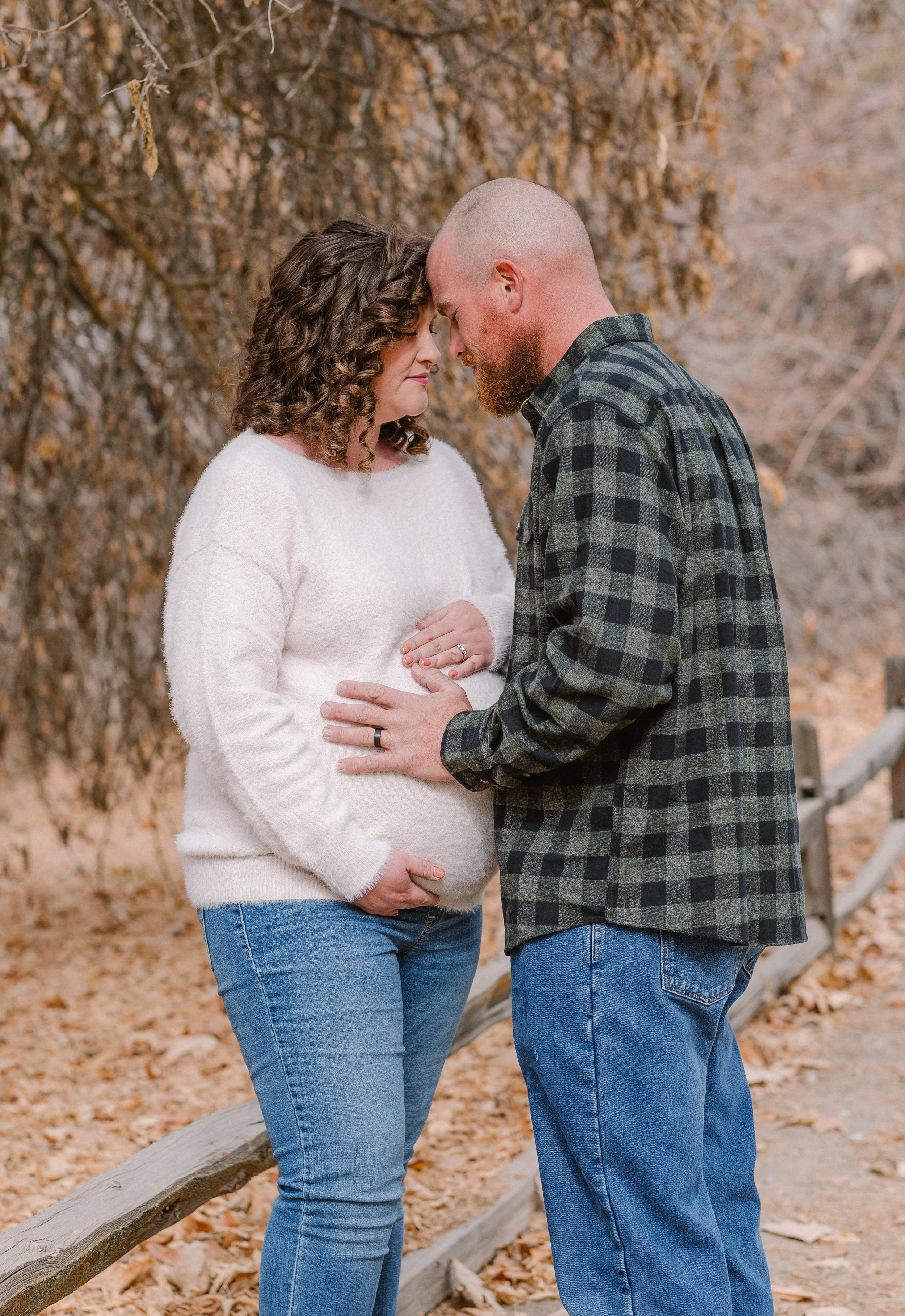 A pregnant woman and a man stand close together outdoors during fall, with trees and fallen leaves, touching foreheads lovingly.
