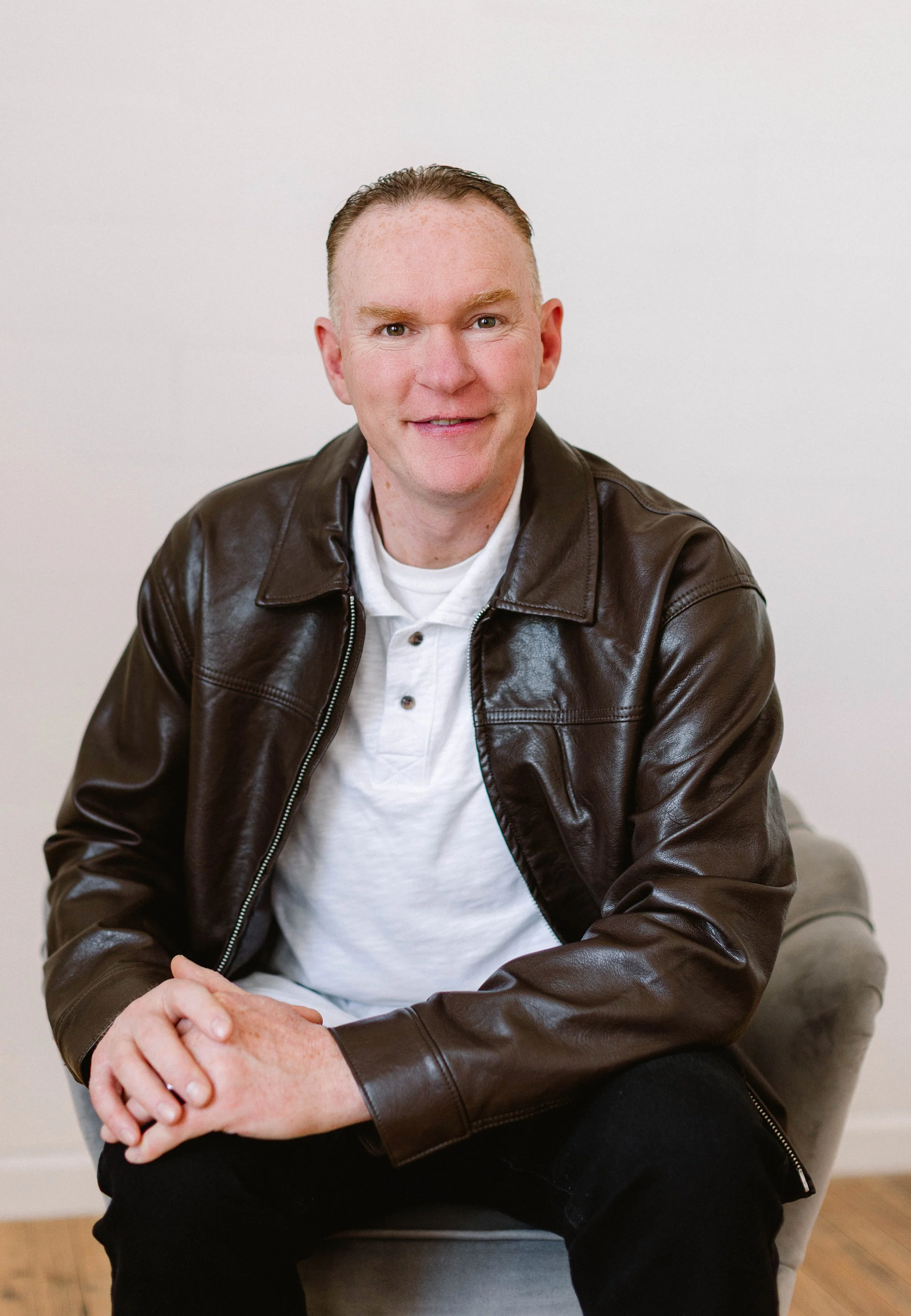 A man with light skin and short reddish hair styled backwards, wearing a brown leather jacket over a white shirt, sitting on a light-colored chair in front of a plain white wall, smiling at the camera.