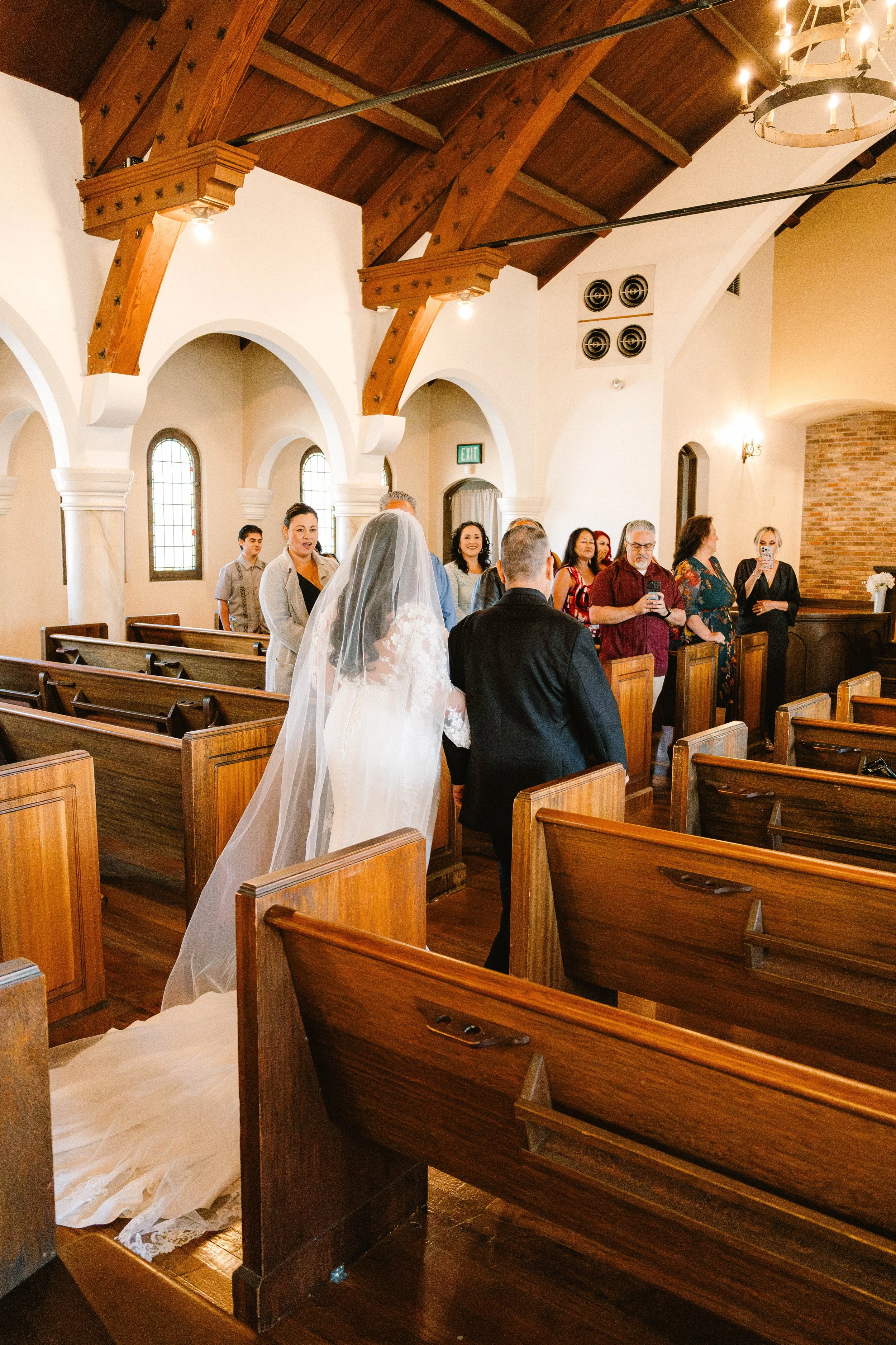 A bride and groom walk down the aisle in a church, surrounded by guests seated in wooden pews.