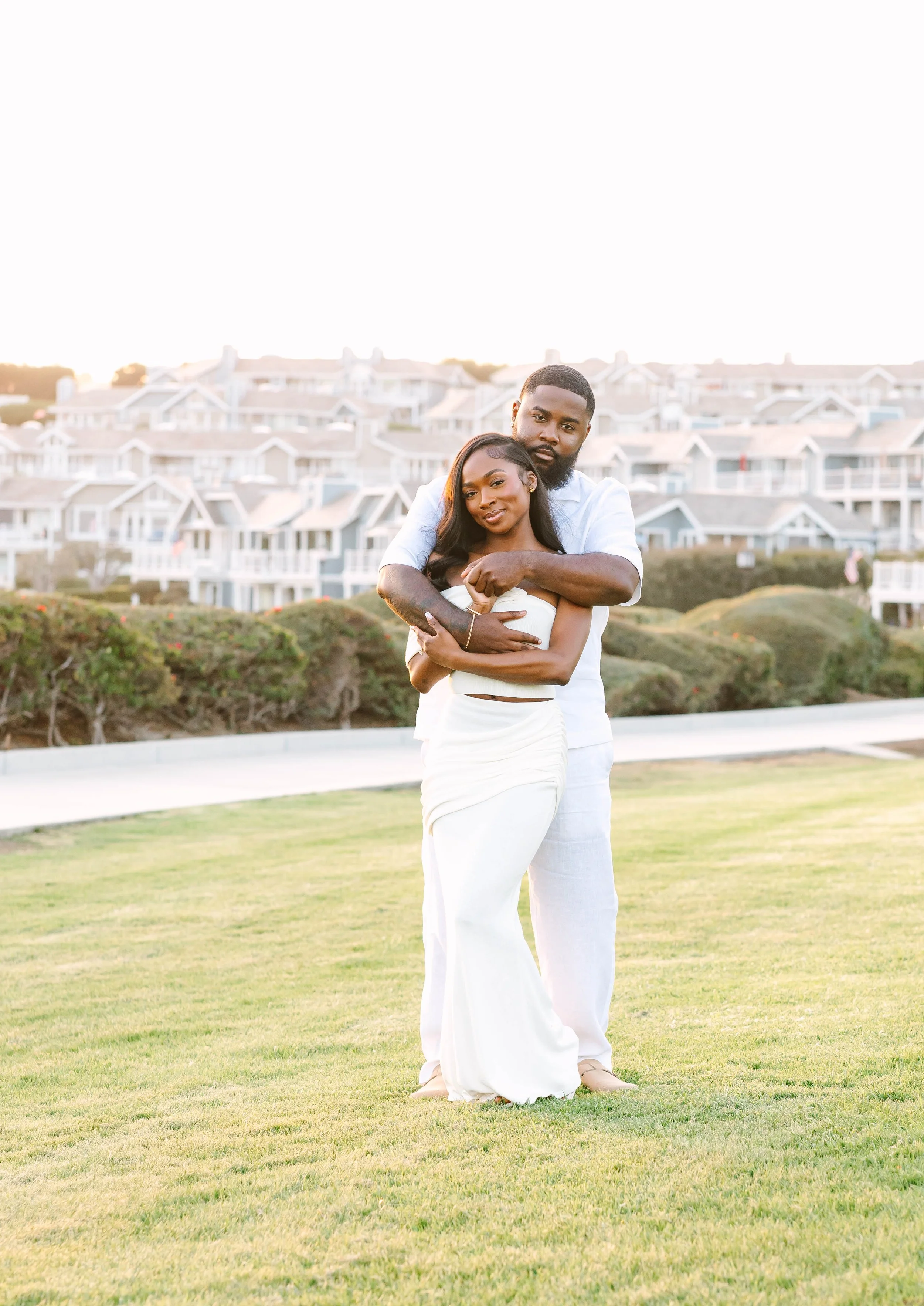 A happy couple standing on a grassy field, embracing each other, with modern houses in the background during sunset.