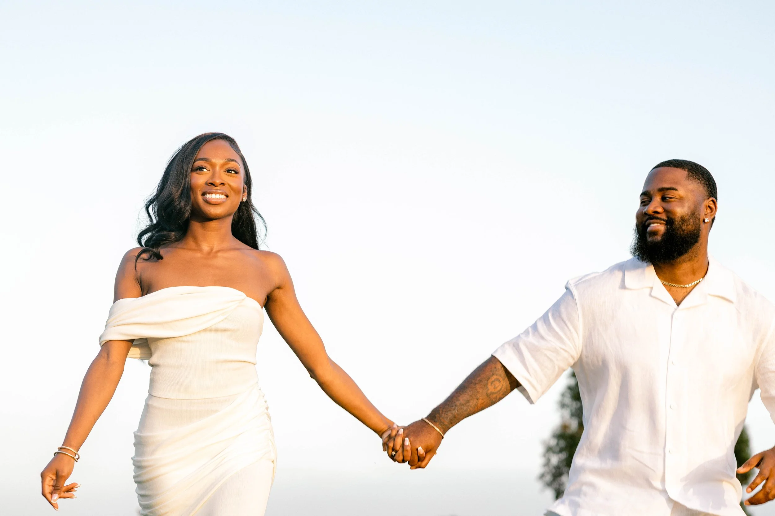 A couple holding hands outdoors, the woman is wearing a strapless white dress and the man is dressed in a white short-sleeved shirt, both smiling and enjoying the moment against a clear sky background.