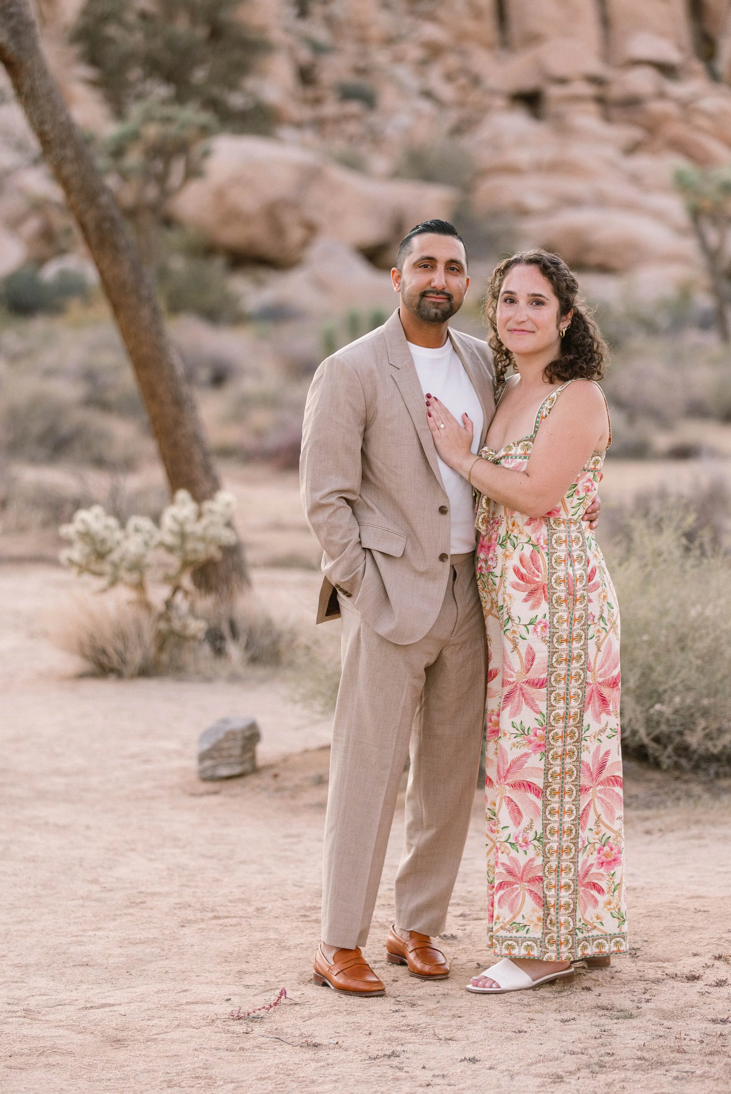 A couple standing close in a desert landscape, with rocks and shrubs in the background. The woman is wearing a sleeveless, floral, wide-leg dress and white sandals, and the man is wearing a beige suit with a white shirt and brown shoes. They are smil