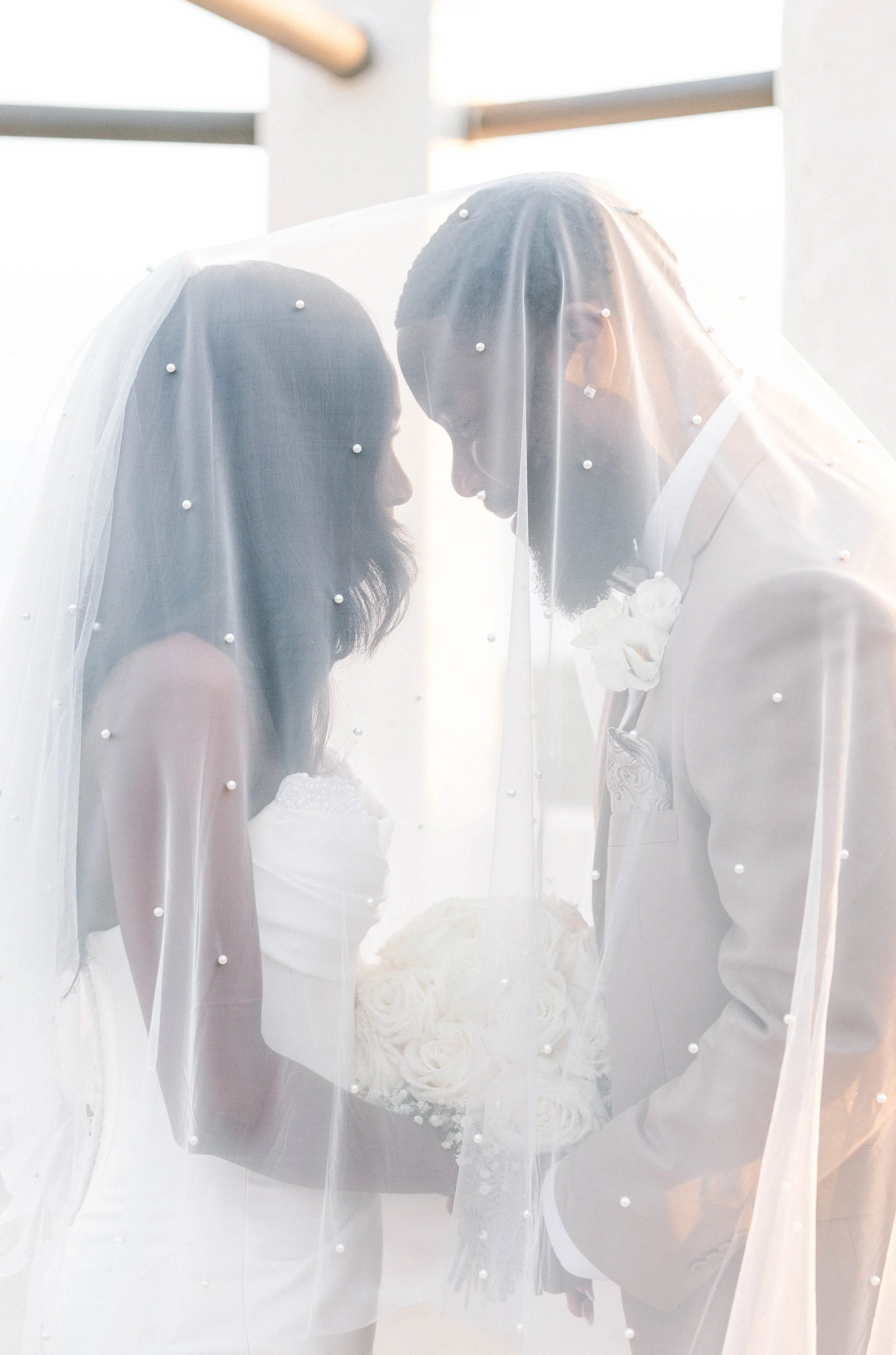 A bride and groom stand close together, their foreheads almost touching, under the bride's veil which is decorated with small pearls. The bride holds a bouquet of white roses, and the groom is dressed in a light-colored suit with a boutonnière. The s