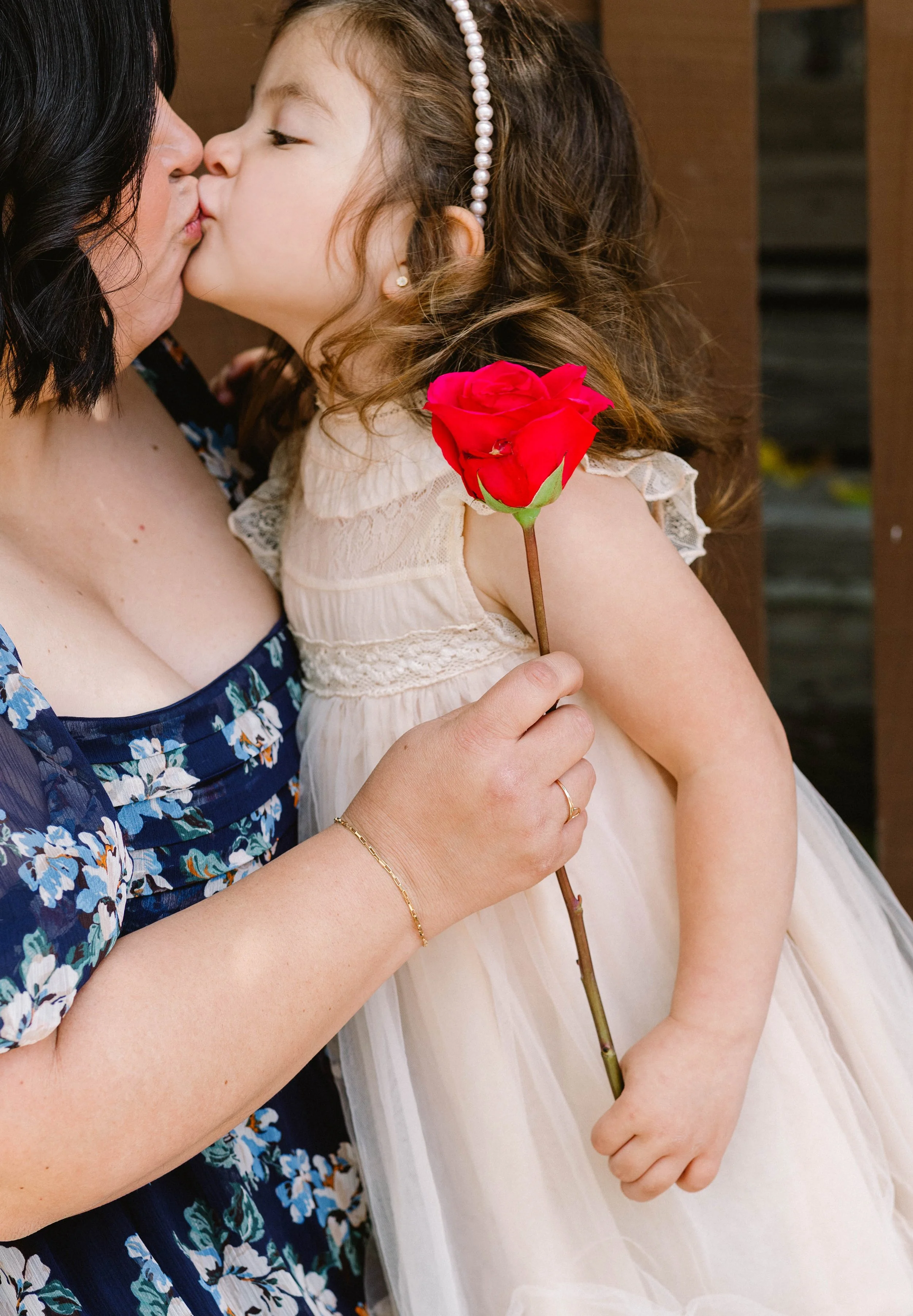 A woman and a young girl share a kiss. The girl holds a red rose and wears a cream dress and a pearl headband. The woman wears a floral dress and a gold bracelet.