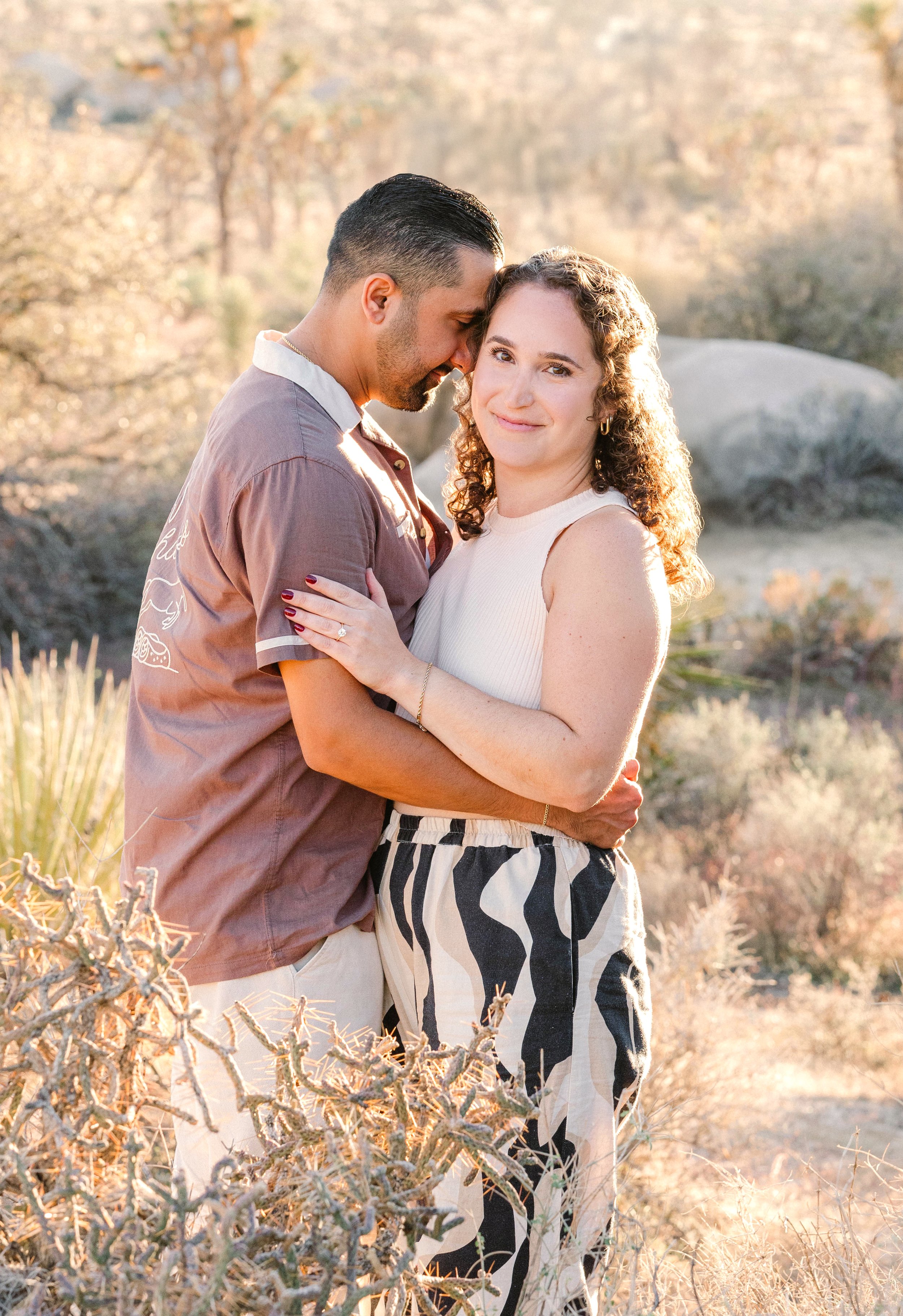 A couple embracing outdoors with desert plants and rocks, during golden hour, sunlight illuminating their faces.