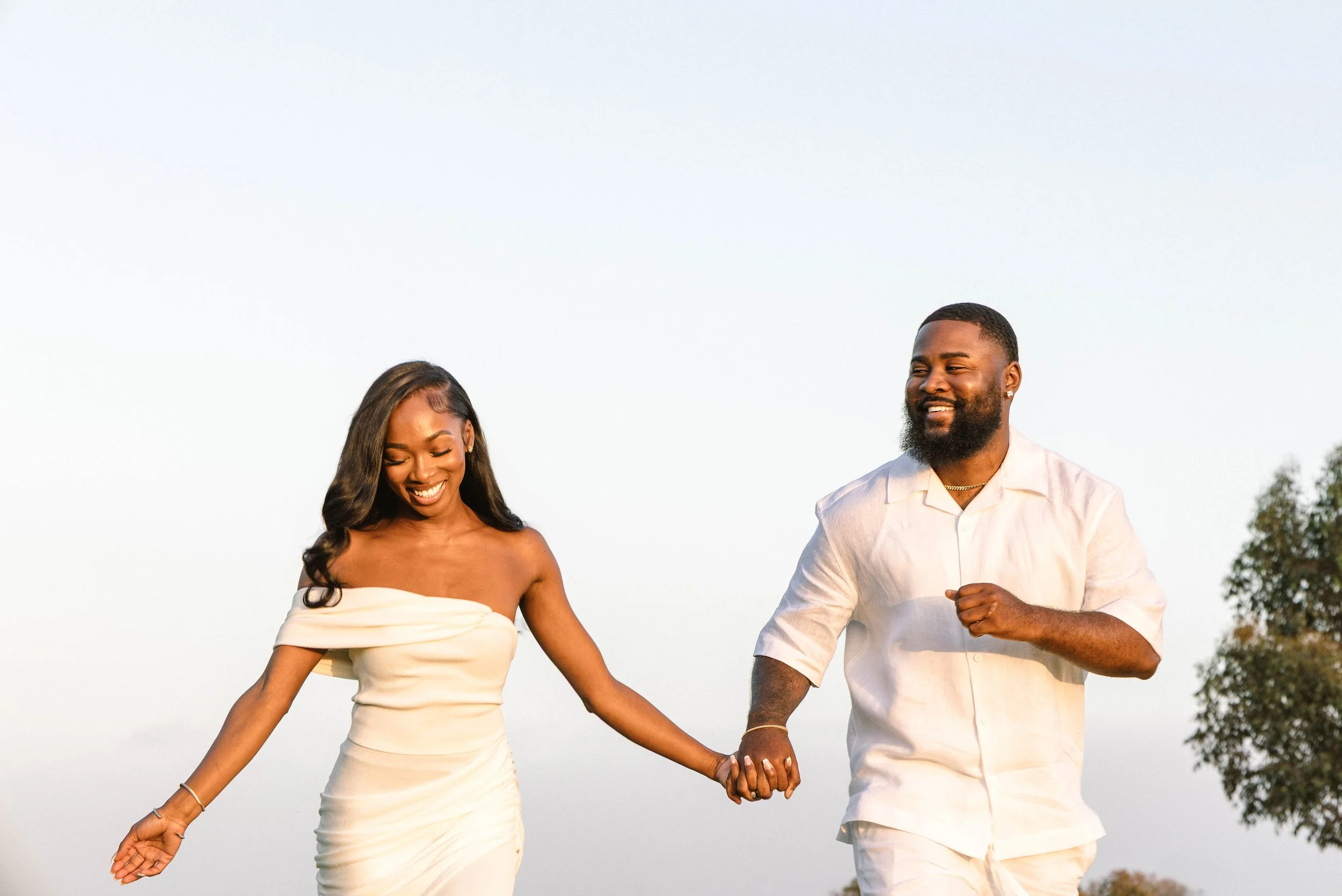 A couple dressed in white, holding hands, smiling and walking outdoors in a natural setting with trees in the background.