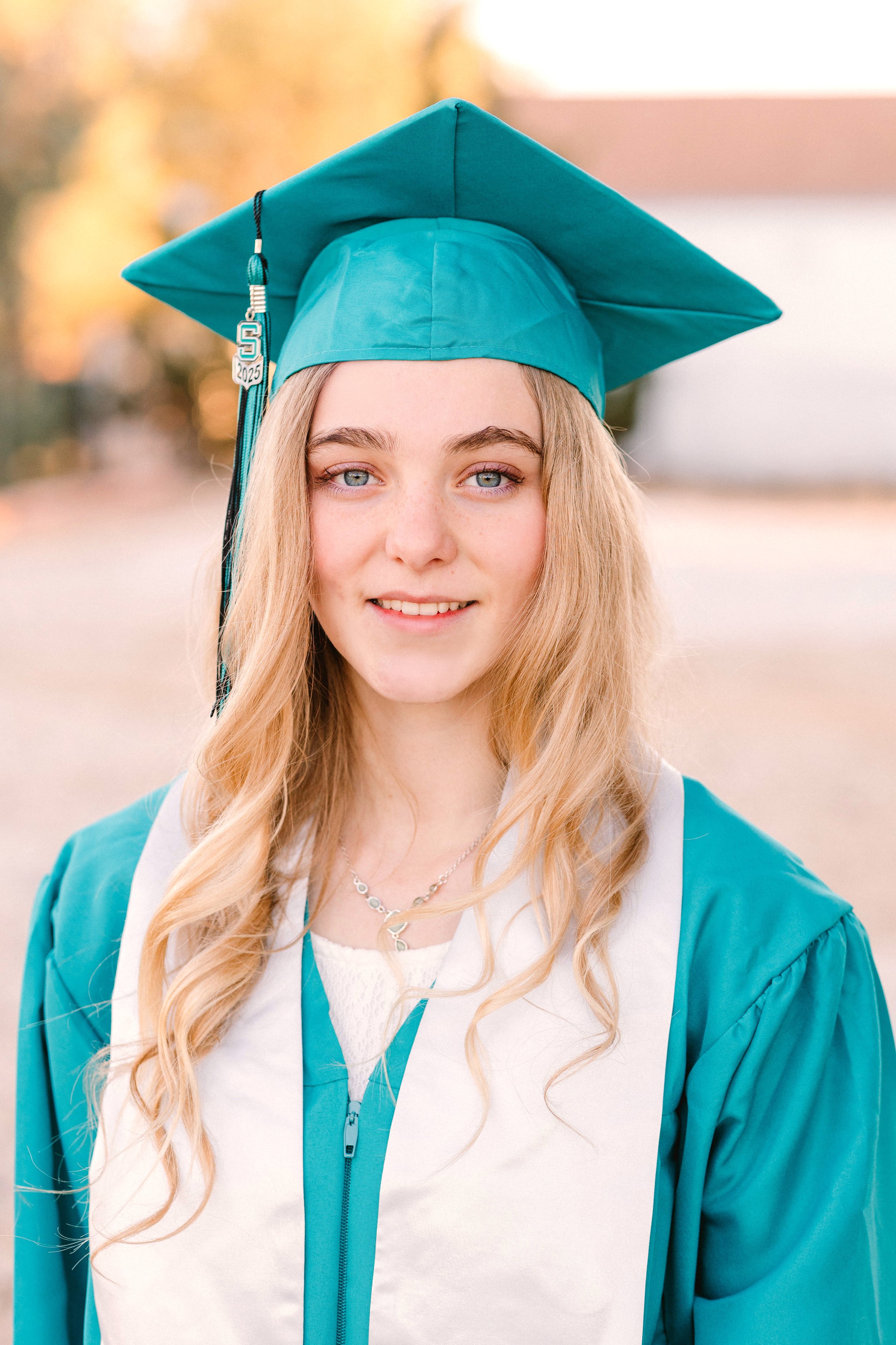 A young woman wearing teal graduation cap and gown, smiling outdoors with blurred autumn trees in the background.
