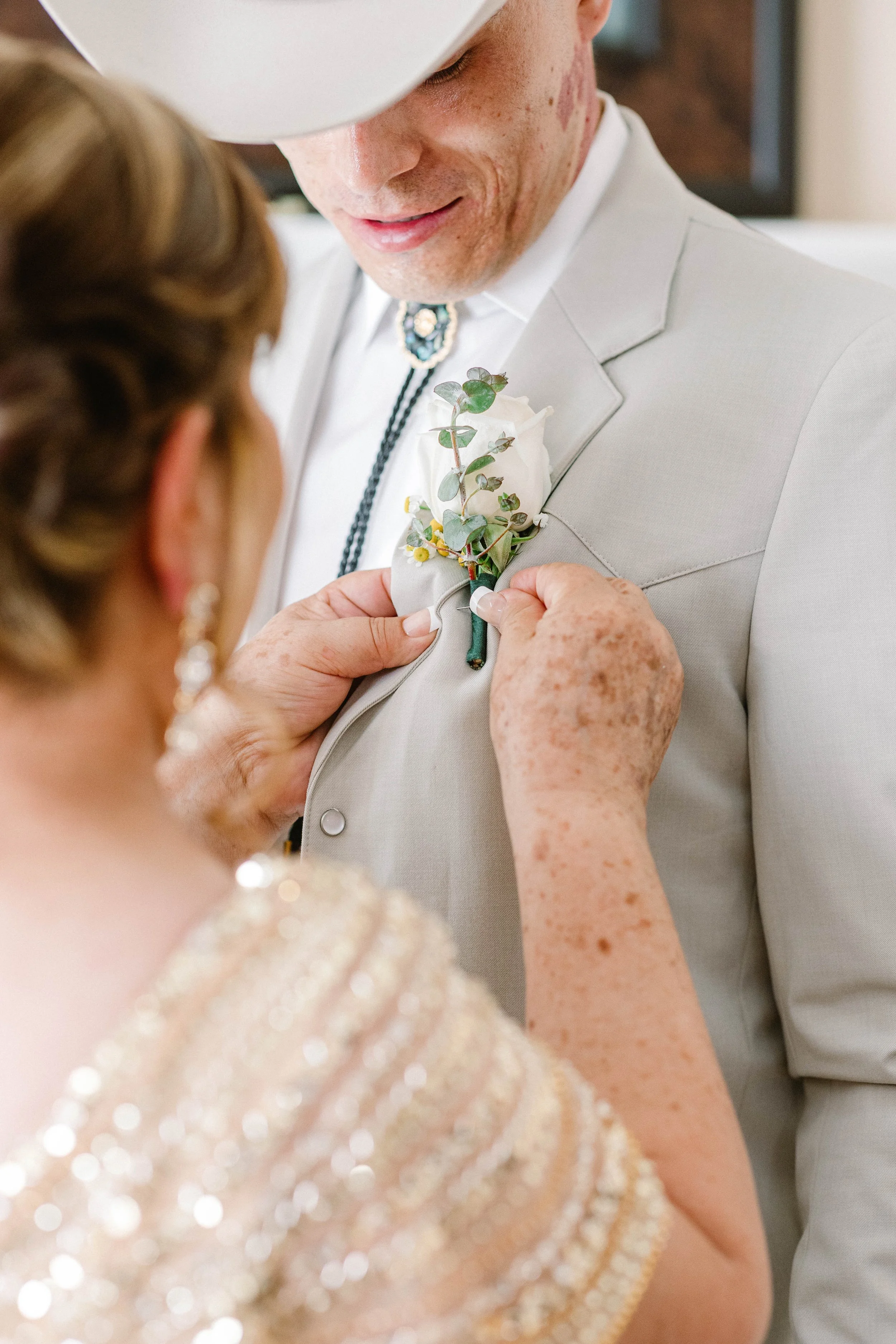 A woman pins a white flower and greenery onto a man's light gray blazer during a wedding or special event.