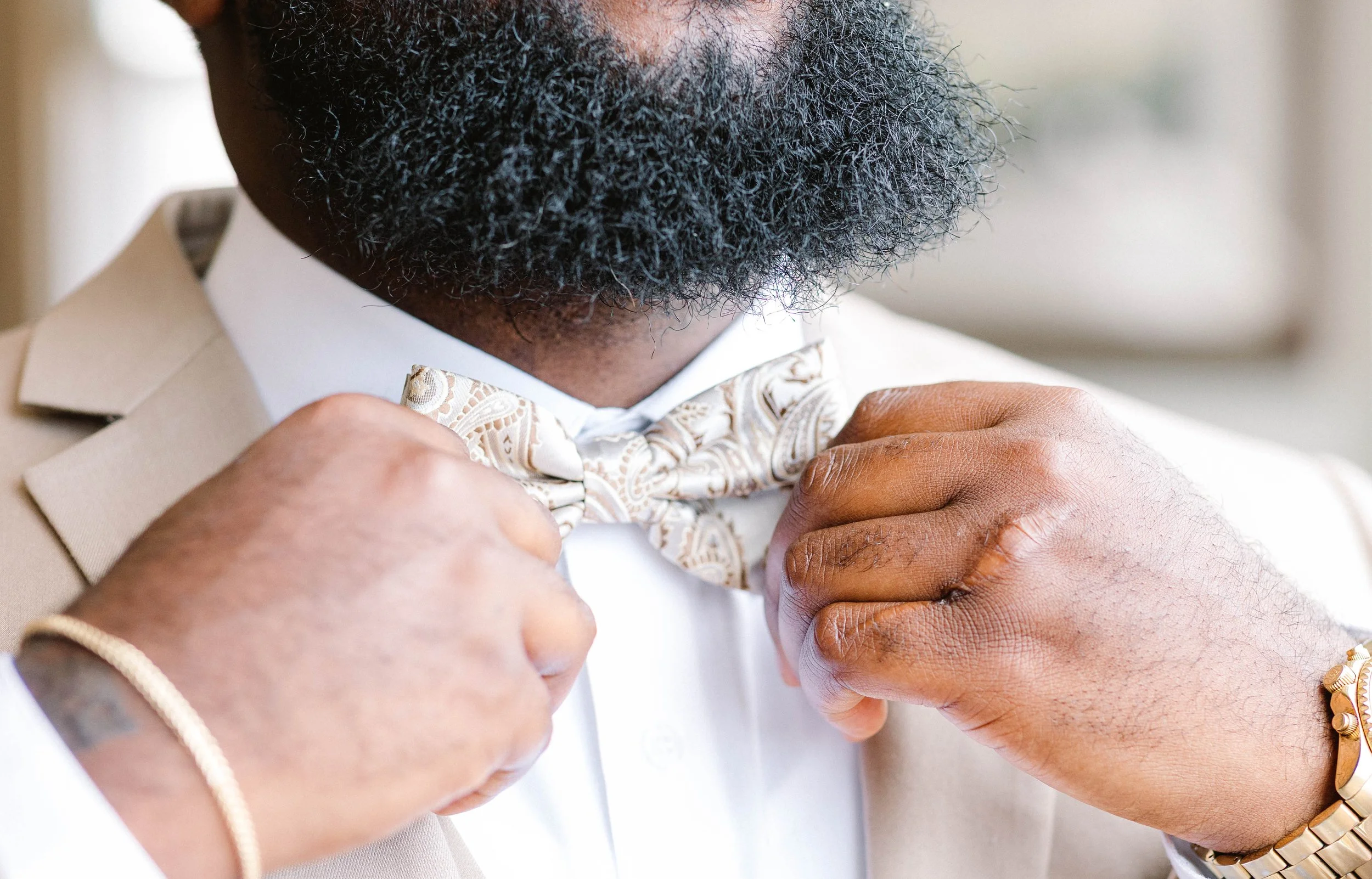 A man adjusting a patterned bow tie on his beige suit and white shirt, wearing a gold watch and bracelet.