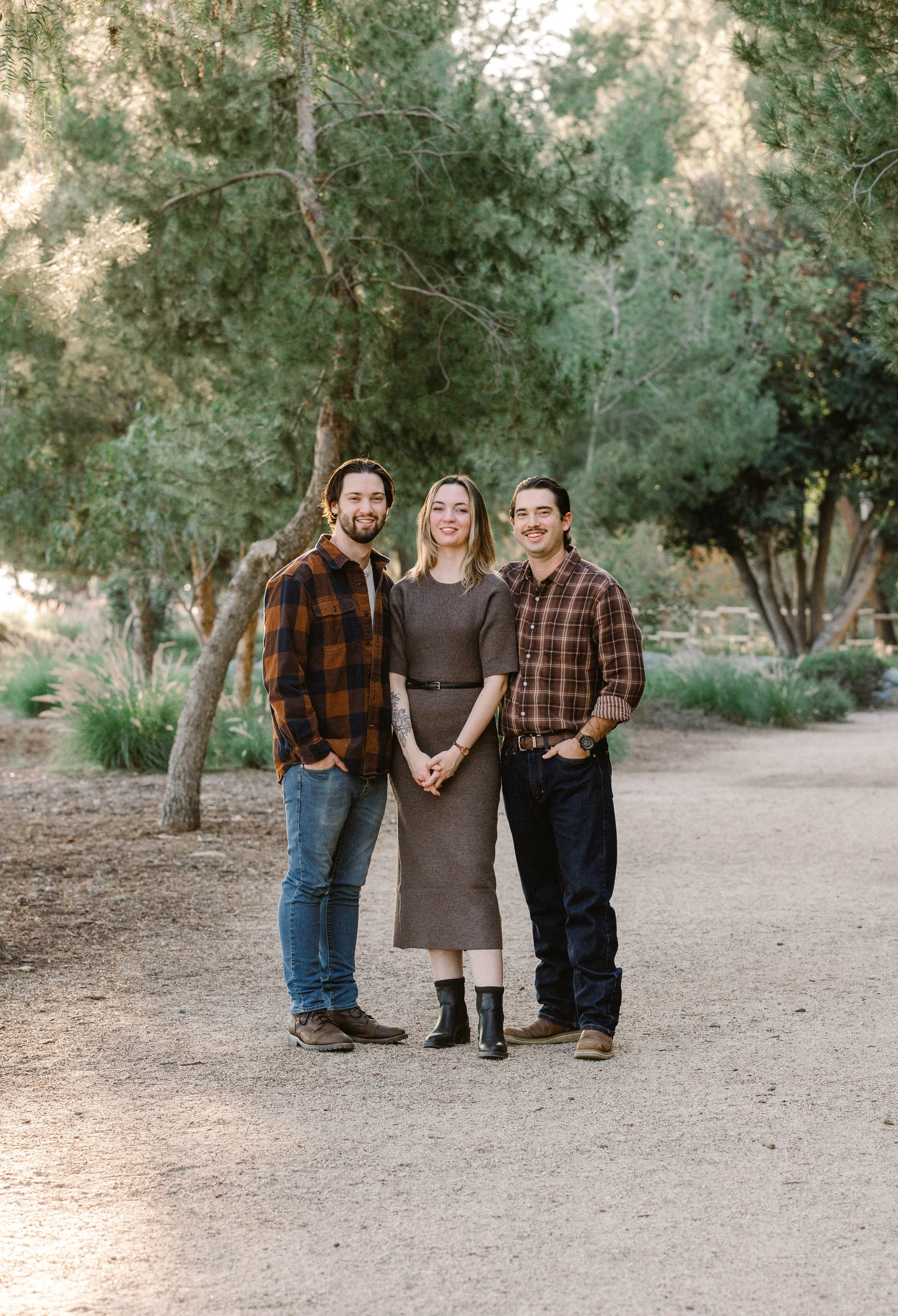 Three people standing outdoors on a dirt pathway, surrounded by green trees and bushes, smiling at the camera during late afternoon.